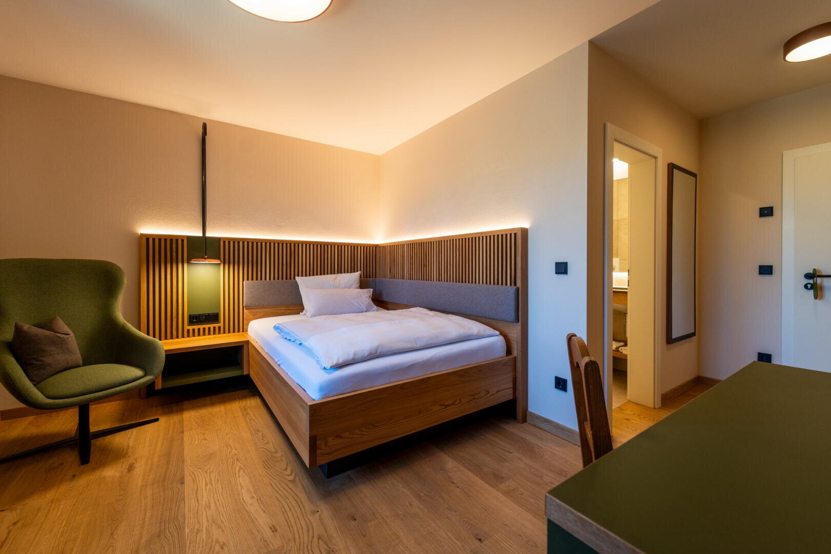 Ein modernes Hotelzimmer mit Einzelbett, grünem Sessel, Holzboden, Schreibtisch und warmer Beleuchtung. Eine offene Tür führt zum Badezimmer. Die Einrichtung ist minimalistisch mit natürlichen Holzakzenten.