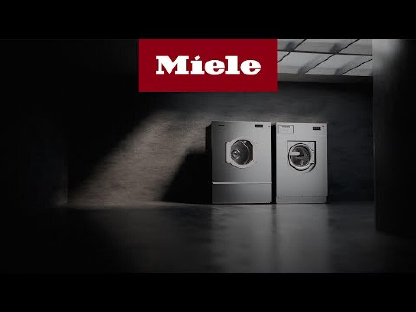 Zwei silberne Miele-Waschmaschinen stehen nebeneinander in einem dunklen, minimalistischen Raum, über dem ein Scheinwerfer angebracht ist. Das Miele-Logo erscheint in weißer Schrift auf rotem Grund oben in der Mitte.