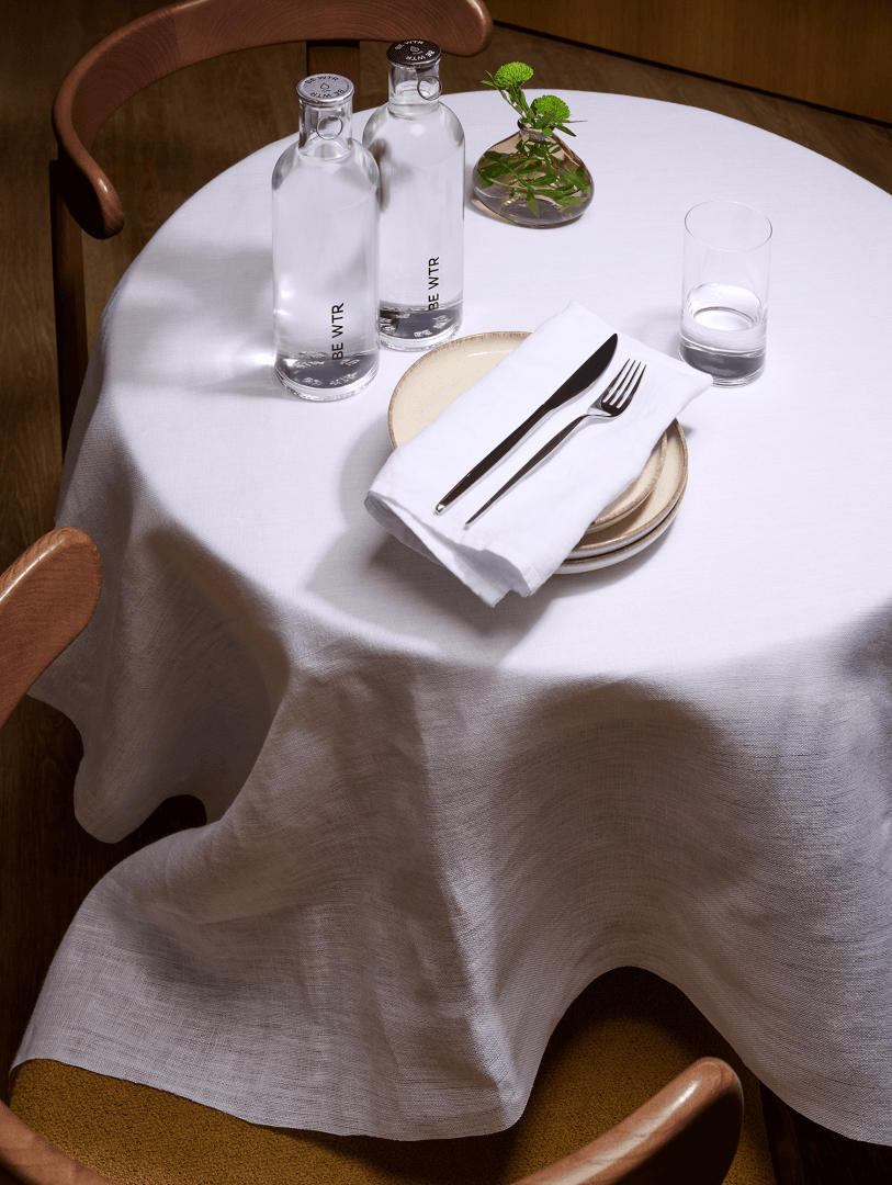 Une table ronde avec une nappe blanche, garnie de deux assiettes, d'une fourchette et d'un couteau sur une serviette pliée, d'un verre d'eau, de deux bouteilles d'eau et d'un petit vase avec des feuilles vertes. Deux chaises en bois sont partiellement visibles.