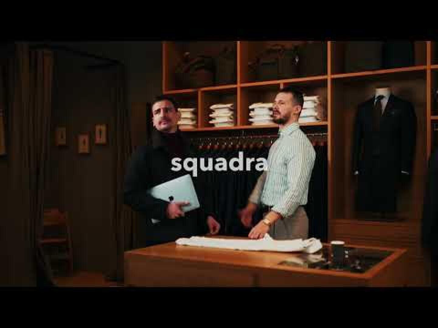 Zwei Männer stehen in einem eleganten Bekleidungsgeschäft, einer hält einen Laptop in der Hand. Im Hintergrund sind Regale mit fein säuberlich angeordneten Artikeln und Anzügen zu sehen. Das Wort Squadra wird quer über das Bild angezeigt.