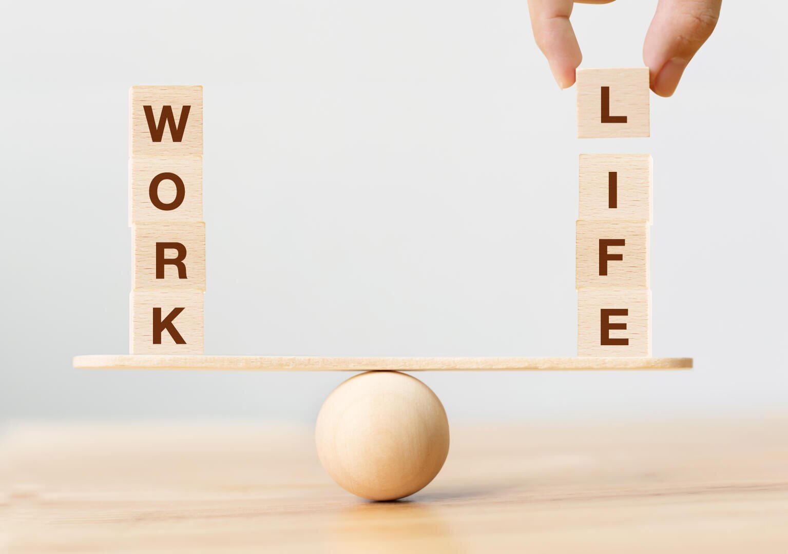 Eine Hand balanciert Holzklötze, auf denen WORK und LIFE geschrieben steht, auf beiden Seiten einer Wippe, die auf einer Holzkugel ruht und das Gleichgewicht zwischen Arbeit und Leben symbolisiert.