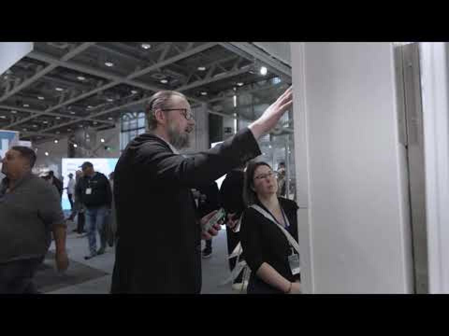 Ein Mann im Anzug gestikuliert in Richtung einer Auslage, während er sich mit einer Frau mit einem Notizblock auf einer überdachten Ausstellung oder Messe unterhält, während im Hintergrund andere Personen und Stände zu sehen sind.