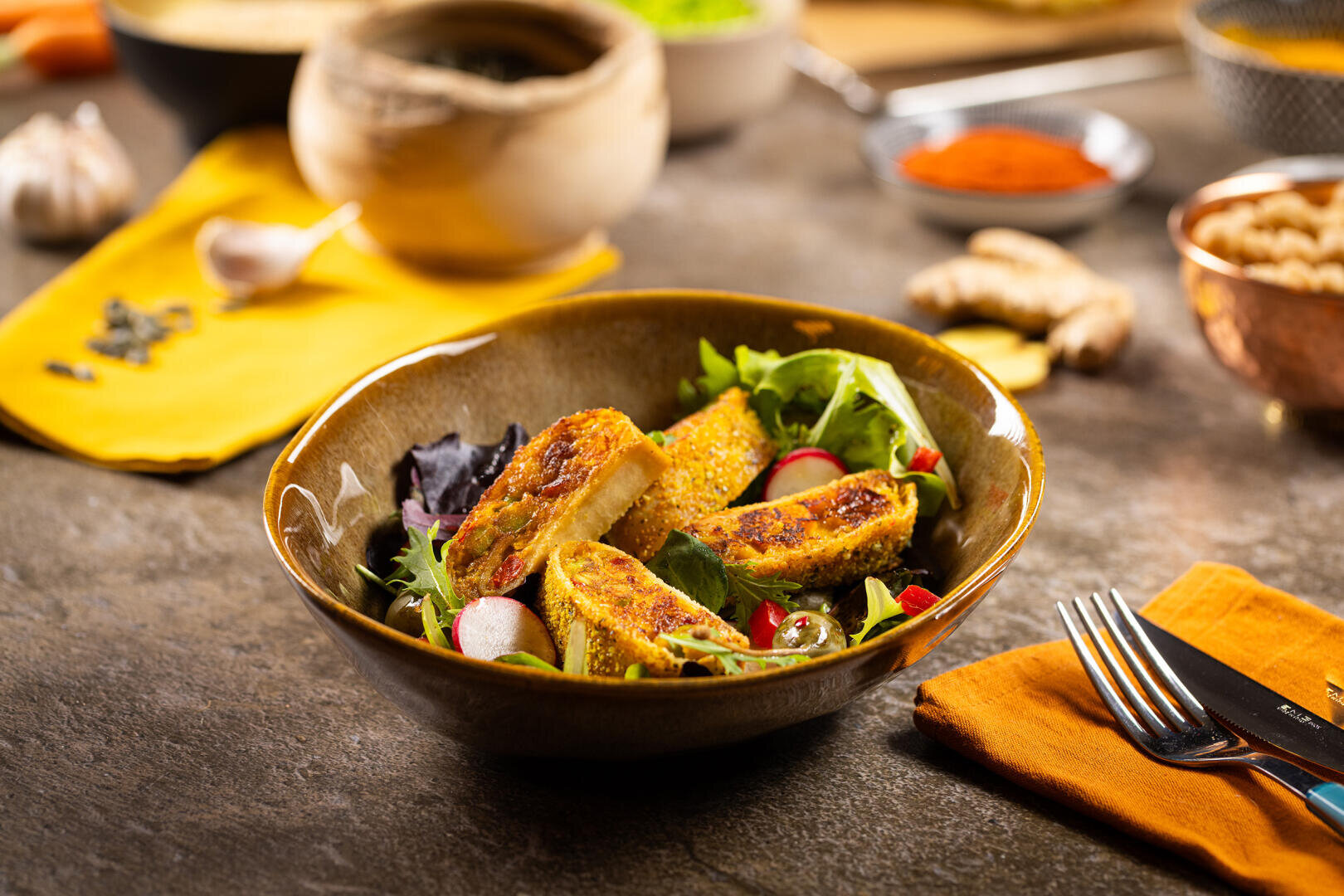 Eine Schüssel mit Salat, belegt mit goldenen, gegrillten Hähnchenscheiben und Radieschen, auf einem rustikalen Tisch mit Utensilien und verschiedenen Zutaten im Hintergrund.