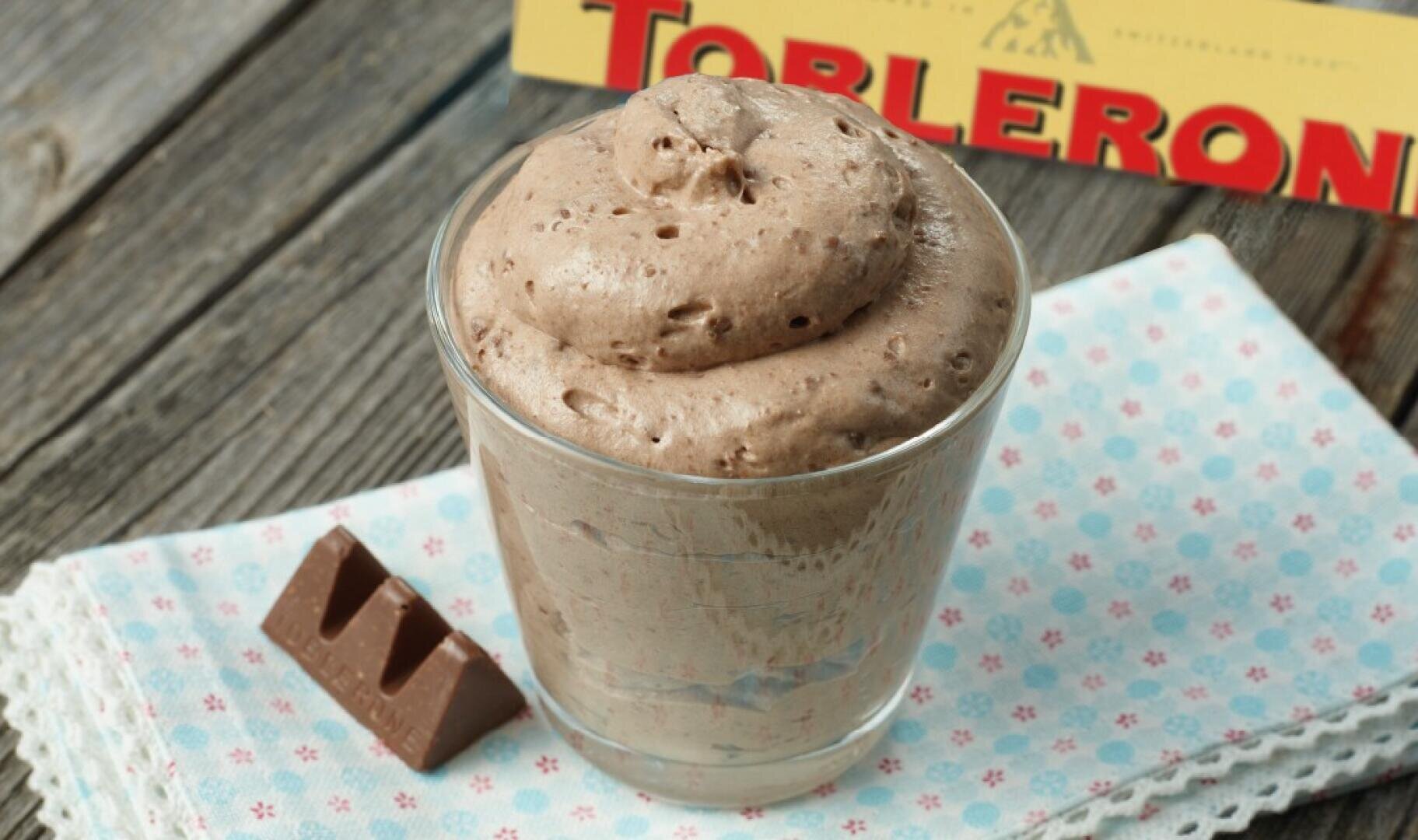 Un verre rempli de mousse au chocolat crémeuse est posé sur une serviette à motifs, à côté d'un morceau de chocolat Toblerone et en arrière-plan une boîte de Toblerone.