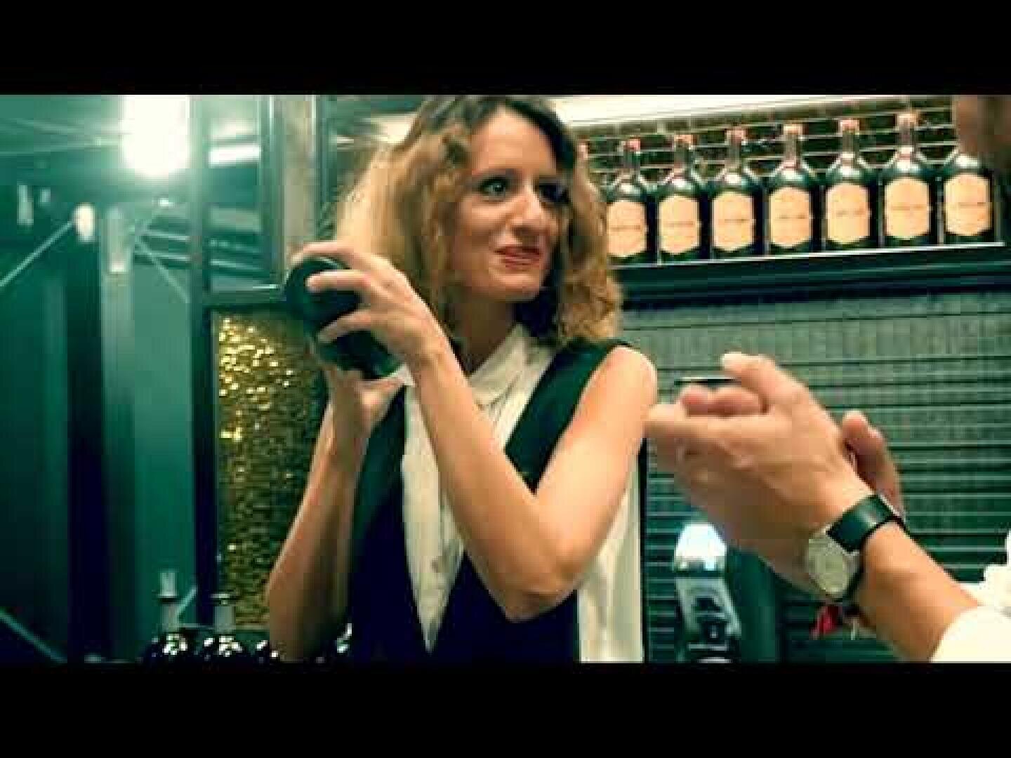 Eine Frau hinter einer Bar schüttelt lächelnd einen Cocktailshaker, während im Hintergrund Flaschen in einem Regal stehen und im Vordergrund die Hände einer anderen Person zu sehen sind.