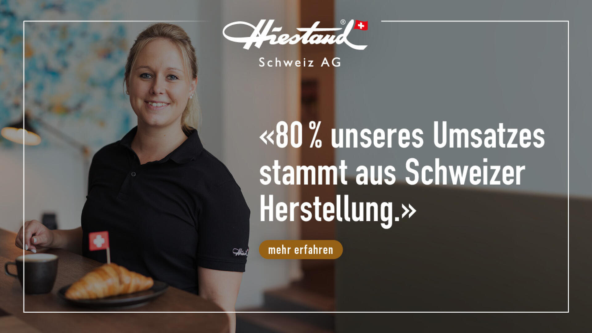 Eine lächelnde Frau in einem schwarzen Poloshirt steht an einem Tisch mit einem Kaffee und einem Croissant, das mit einer Schweizer Flagge verziert ist. Der Text lautet: 80% unseres Umsatzes stammt aus Schweizer Herstellung.