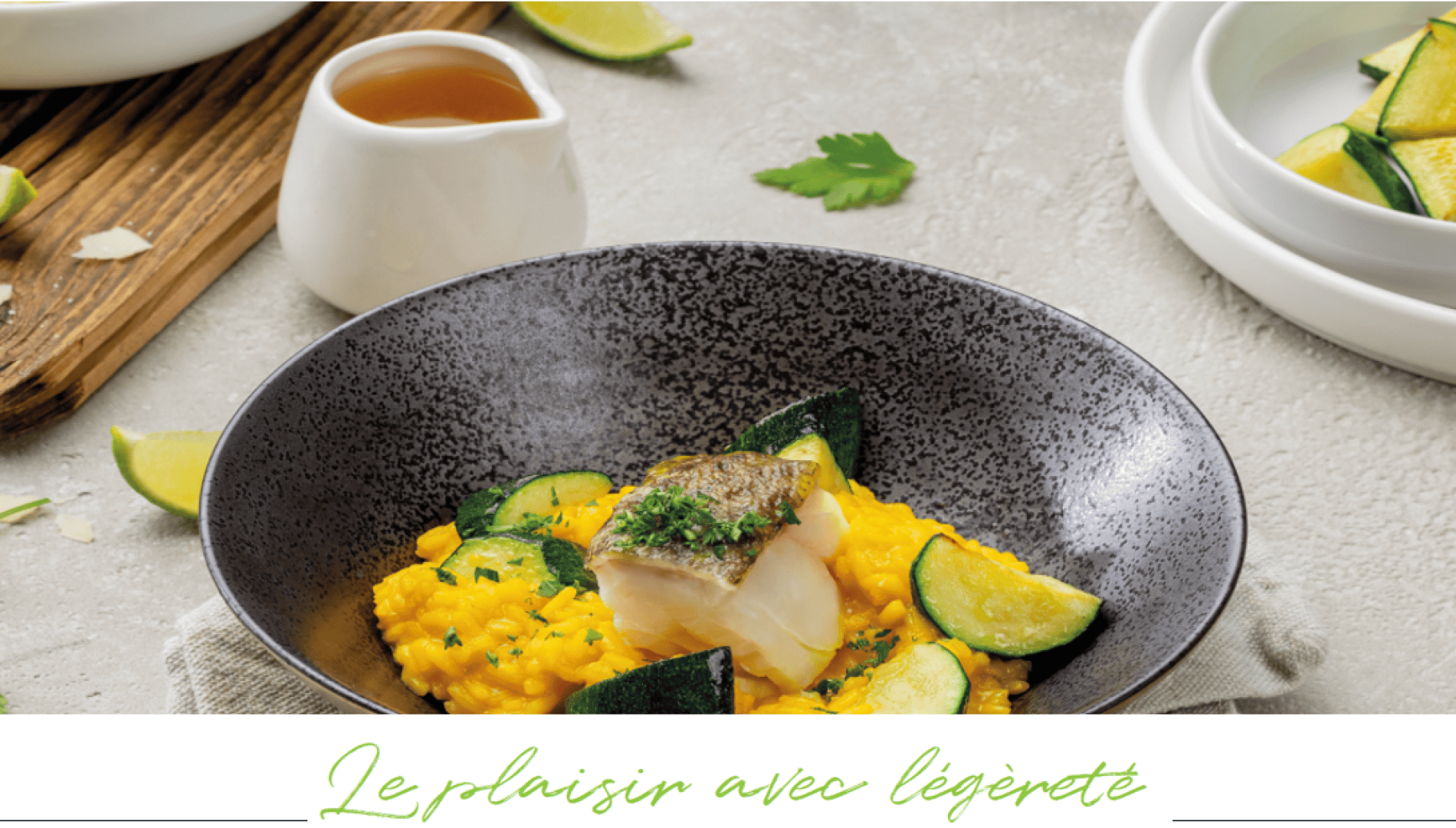 Un bol de risotto garni de courgettes et d'un morceau de poisson, agrémenté d'herbes. Un pot de sauce, des quartiers de citron vert et du persil sont placés autour du bol. Le texte est le suivant : Le plaisir avec légèreté.