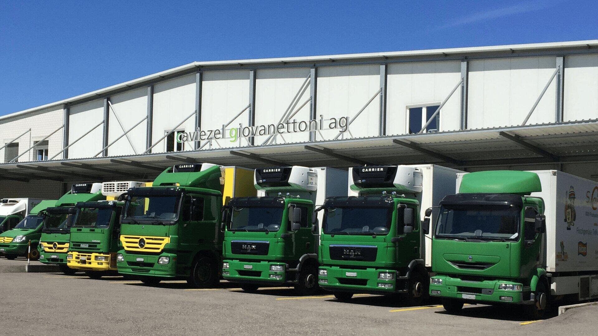 Eine Reihe grüner und gelber Lieferwagen ist an einem sonnigen Tag vor einem Lagerhaus geparkt. Das Gebäude trägt ein Schild mit der Aufschrift giovanettoni ag über den Toren der Laderampe.