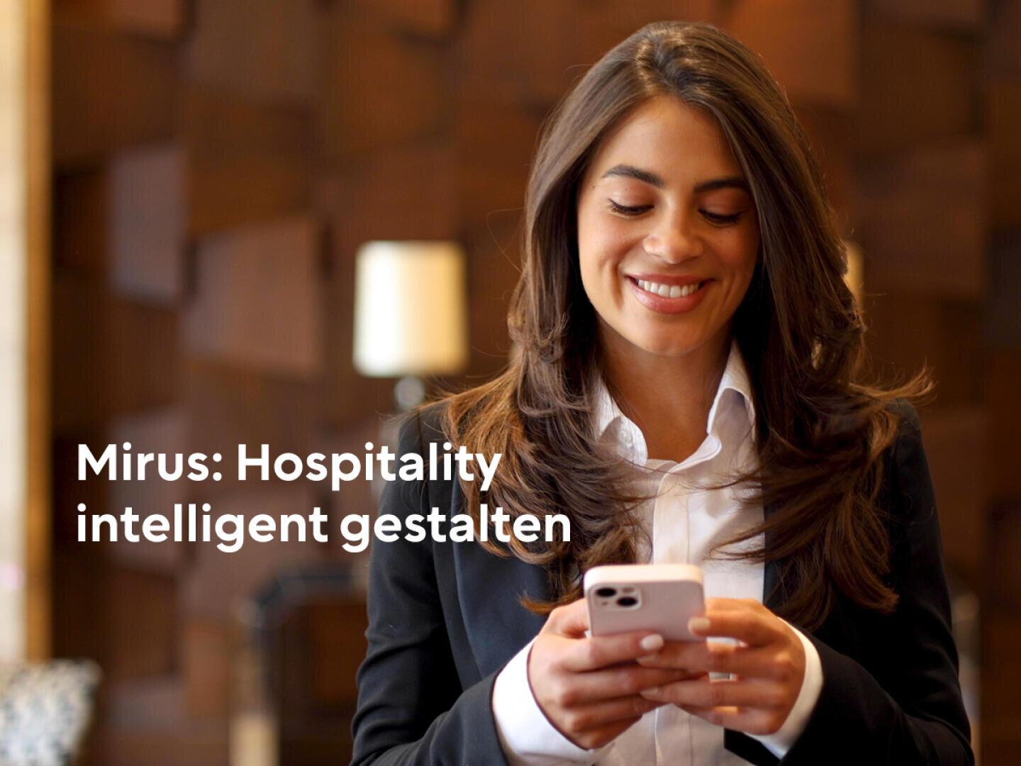 Une femme souriante en tenue professionnelle utilise un smartphone. L'arrière-plan est flou et laisse apparaître un mur marron à motifs. Le texte sur l'image est le suivant : Mirus : Concevoir l'hospitalité intelligemment.