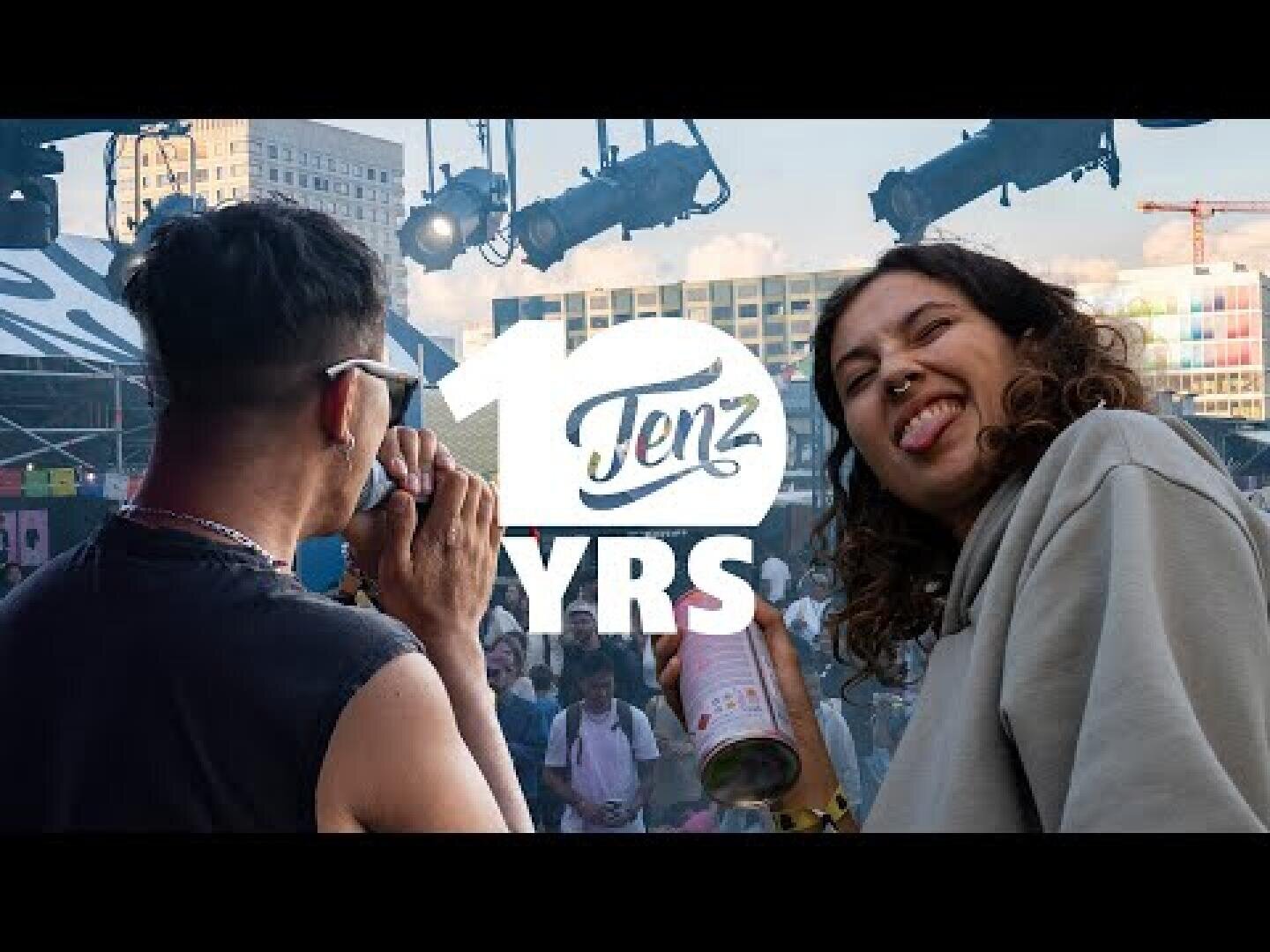 Zwei Menschen stehen vor einer Festivalbühne; einer hält eine Kamera, der andere lächelt und streckt die Zunge heraus. In der Mitte ist der Text 12 TenZ YRS eingeblendet. Im Hintergrund sind Bühnenlichter und eine Stadtlandschaft zu sehen.
