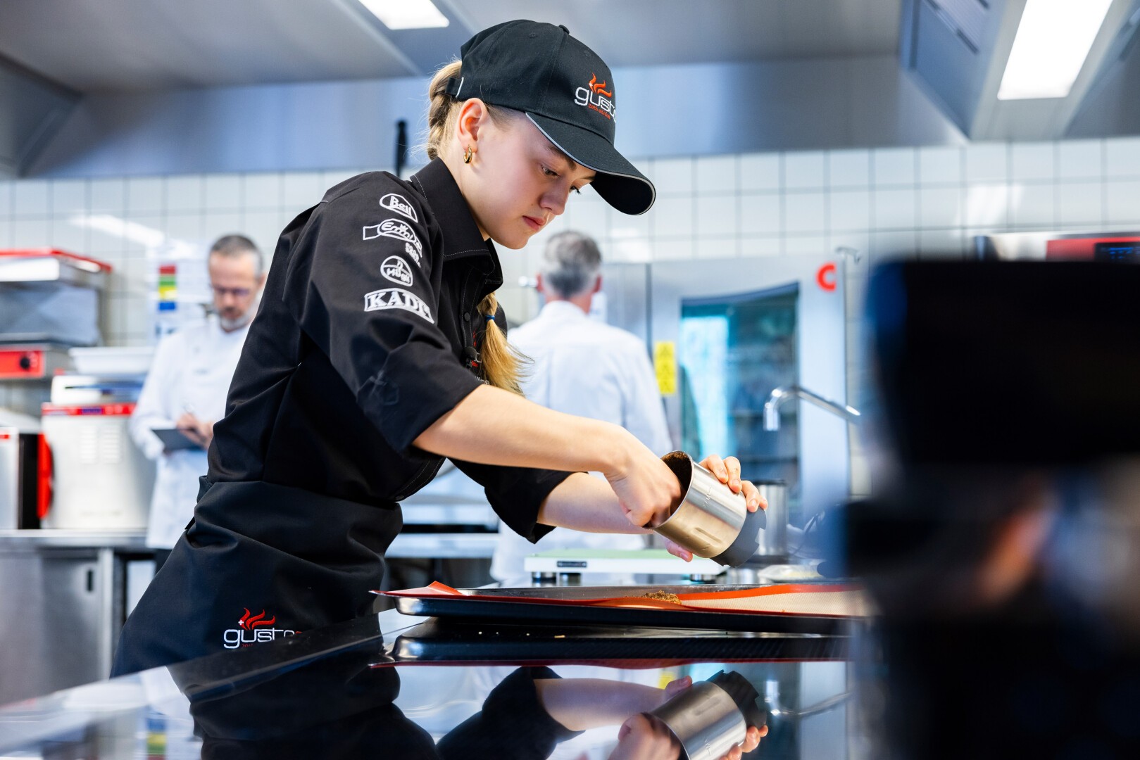 Un chef en uniforme noir et casquette verse du liquide d'un petit pichet en métal sur un plat dans une cuisine moderne et professionnelle, avec d'autres chefs et des appareils en acier inoxydable en arrière-plan.