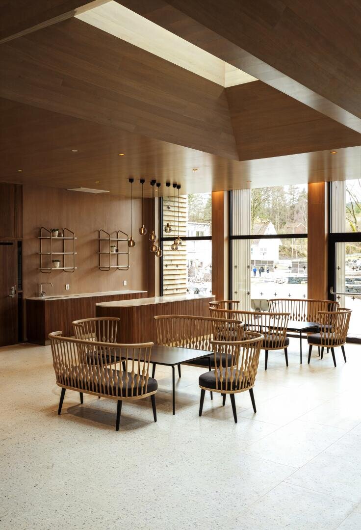 Eine moderne Lounge mit Holzvertäfelung, großen Fenstern, runden Tischen und Rattanstühlen mit schwarzen Kissen, die unter Hängelampen stehen. Natürliches Licht erfüllt den geräumigen, minimalistischen Innenraum.