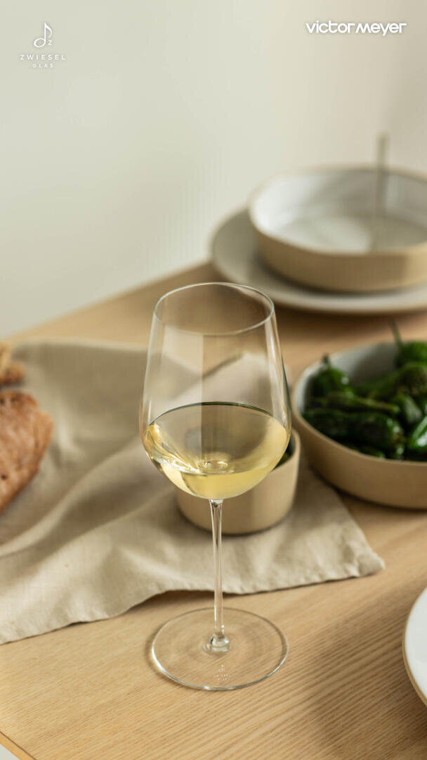 Ein Glas Weißwein steht auf einem Holztisch, der mit Tellern, einer Schale mit grünem Paprika, Brot und einer beigen Serviette gedeckt ist. Die Szene ist hell und einladend, was auf ein zwangloses Essen hindeutet.
