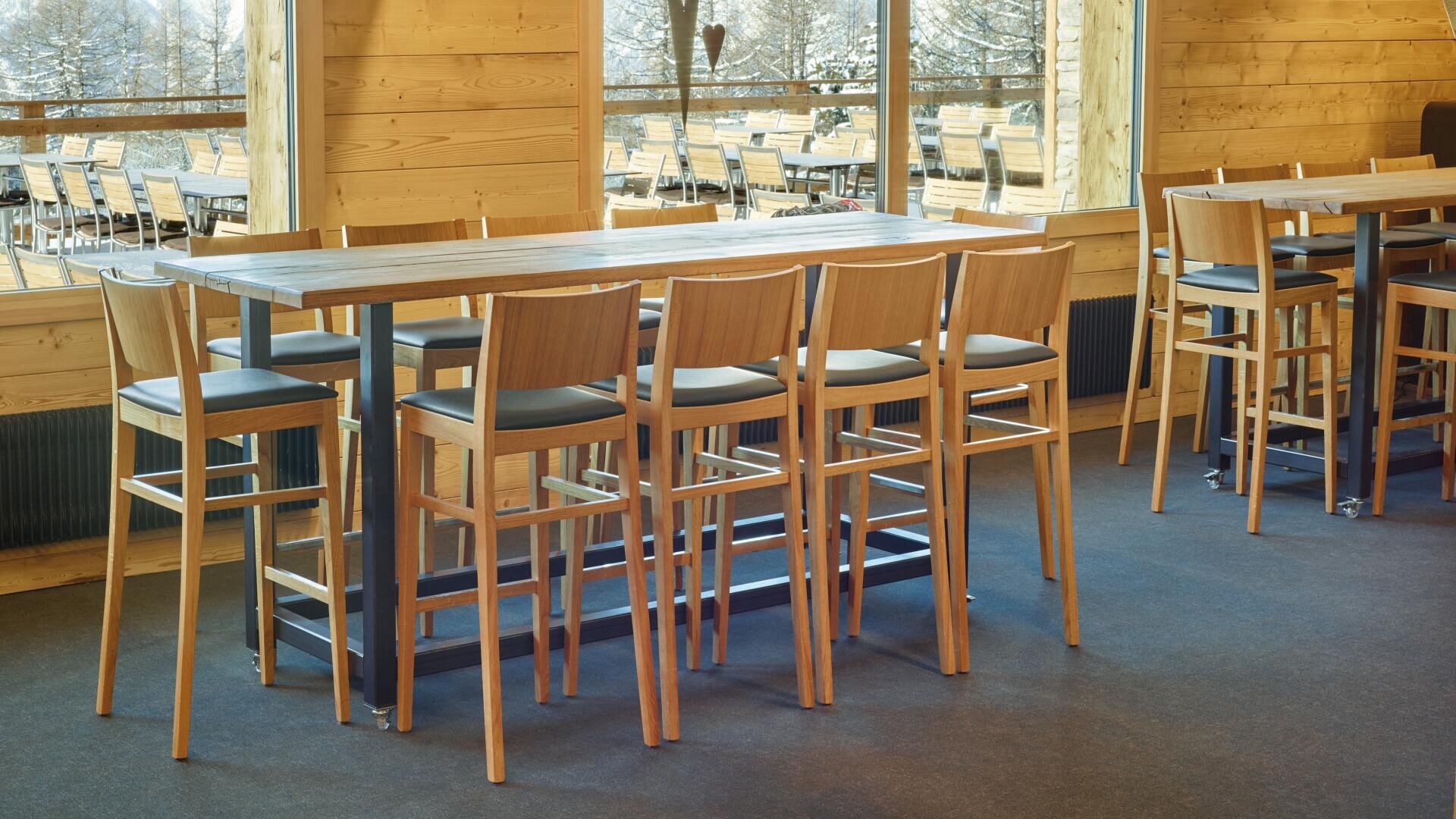 Une salle à manger moderne avec de hautes tables en bois et des chaises hautes assorties, placées près de grandes fenêtres qui permettent de s'asseoir à l'extérieur et offrent une vue pittoresque sur les arbres. L'intérieur présente des murs en bois et une lumière naturelle.