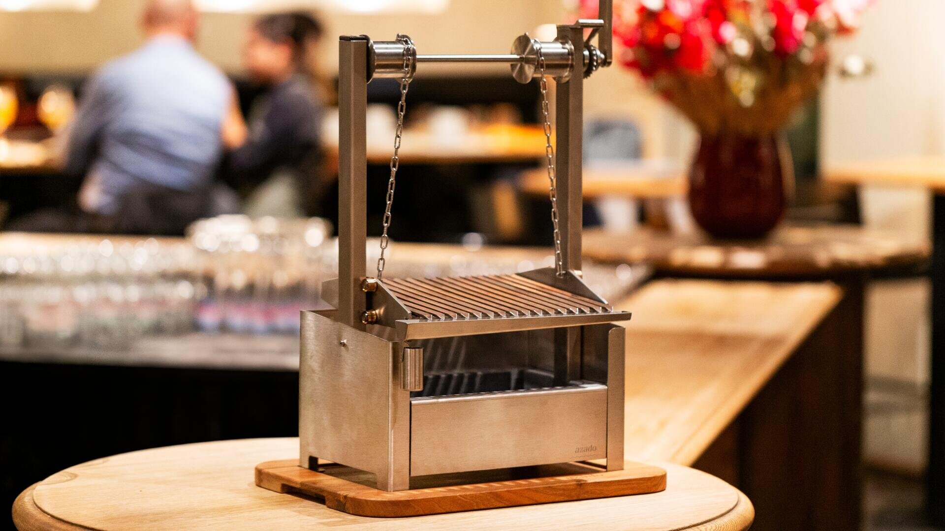 Ein kleiner Tischgrill aus rostfreiem Stahl mit einem hölzernen Untergestell und einem verstellbaren Rost steht auf einem runden Holztisch in einem Restaurant, mit unscharfen Gästen und Blumen im Hintergrund.