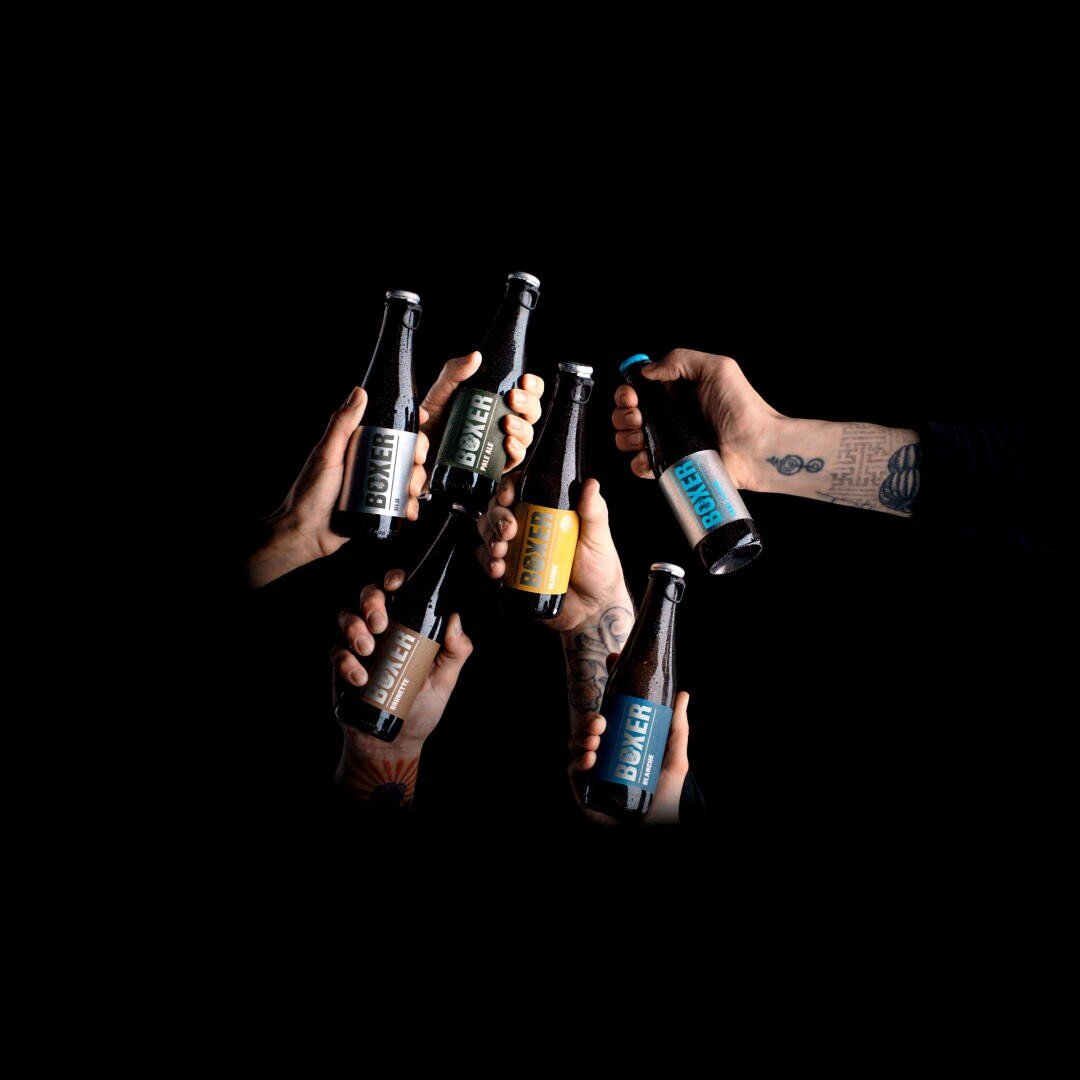 Sechs Hände halten verschiedene Bierflaschen mit bunten Etiketten vor einem schwarzen Hintergrund. Eine Hand zeigt Tätowierungen auf dem Unterarm, und auf jedem Flaschenetikett steht Bôler.