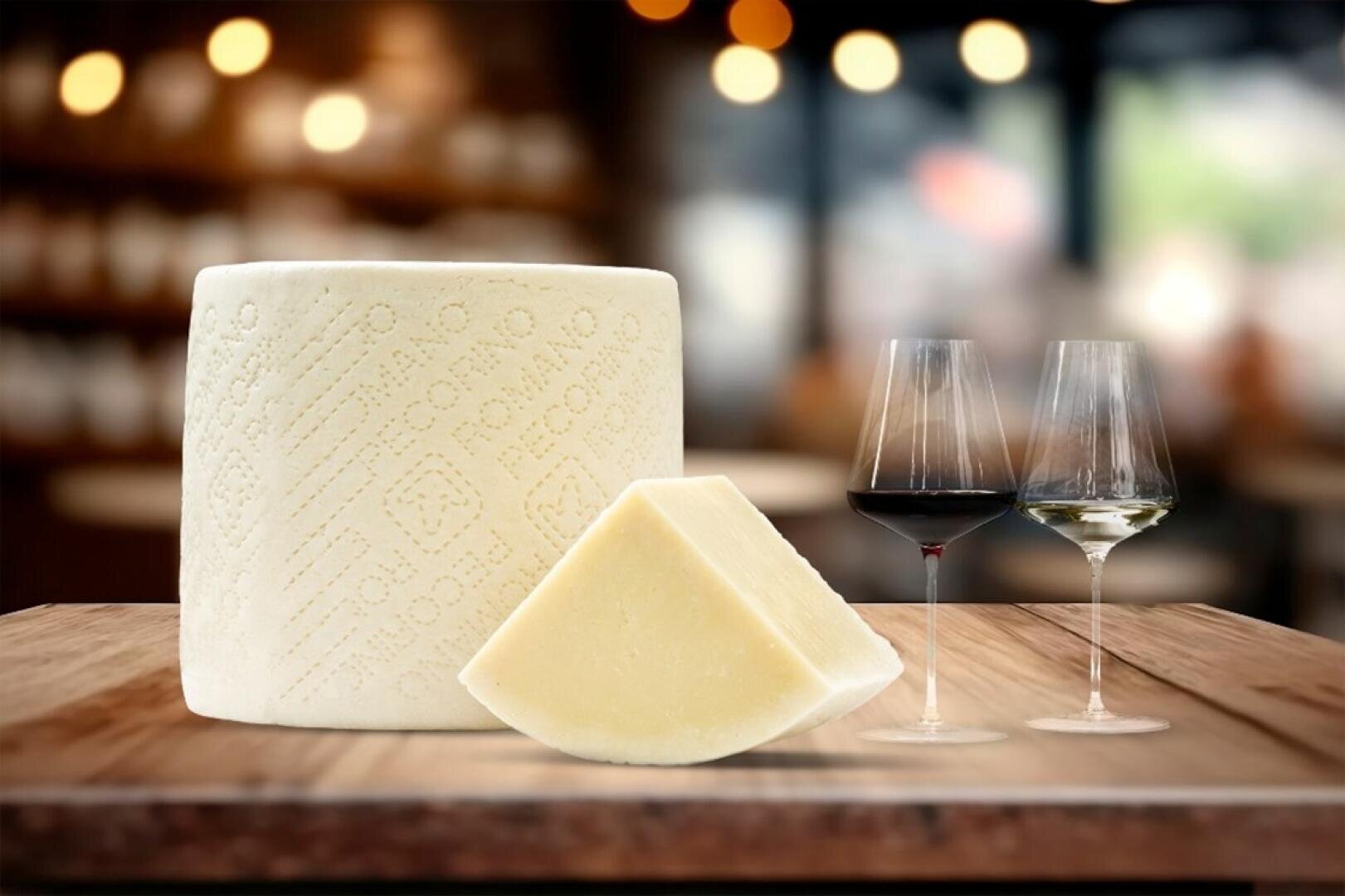 Une meule de fromage de brebis (pecorino romano) avec un morceau découpé est posée sur un support en bois à côté de deux verres de vin, un rouge et un blanc, avec un arrière-plan de restaurant légèrement flou.