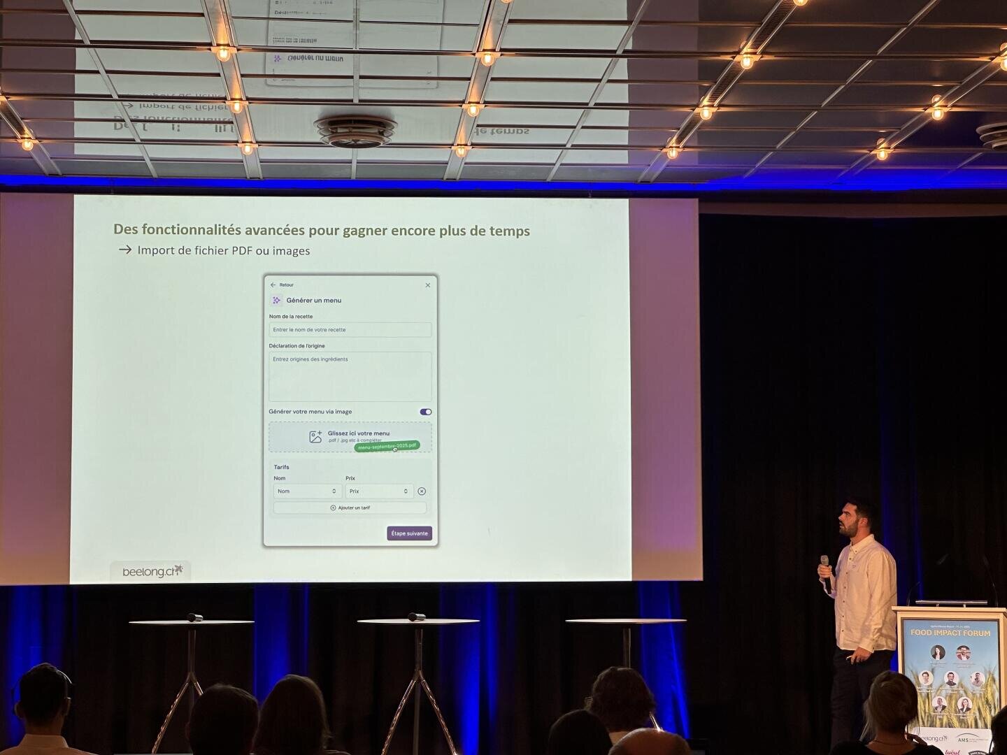 Un présentateur se tient sur scène près d'un grand écran affichant une diapositive en français sur les fonctions avancées permettant de gagner du temps, montrant une interface pour l'importation de fichiers PDF ou d'images. Les membres de l'auditoire sont assis au premier plan.