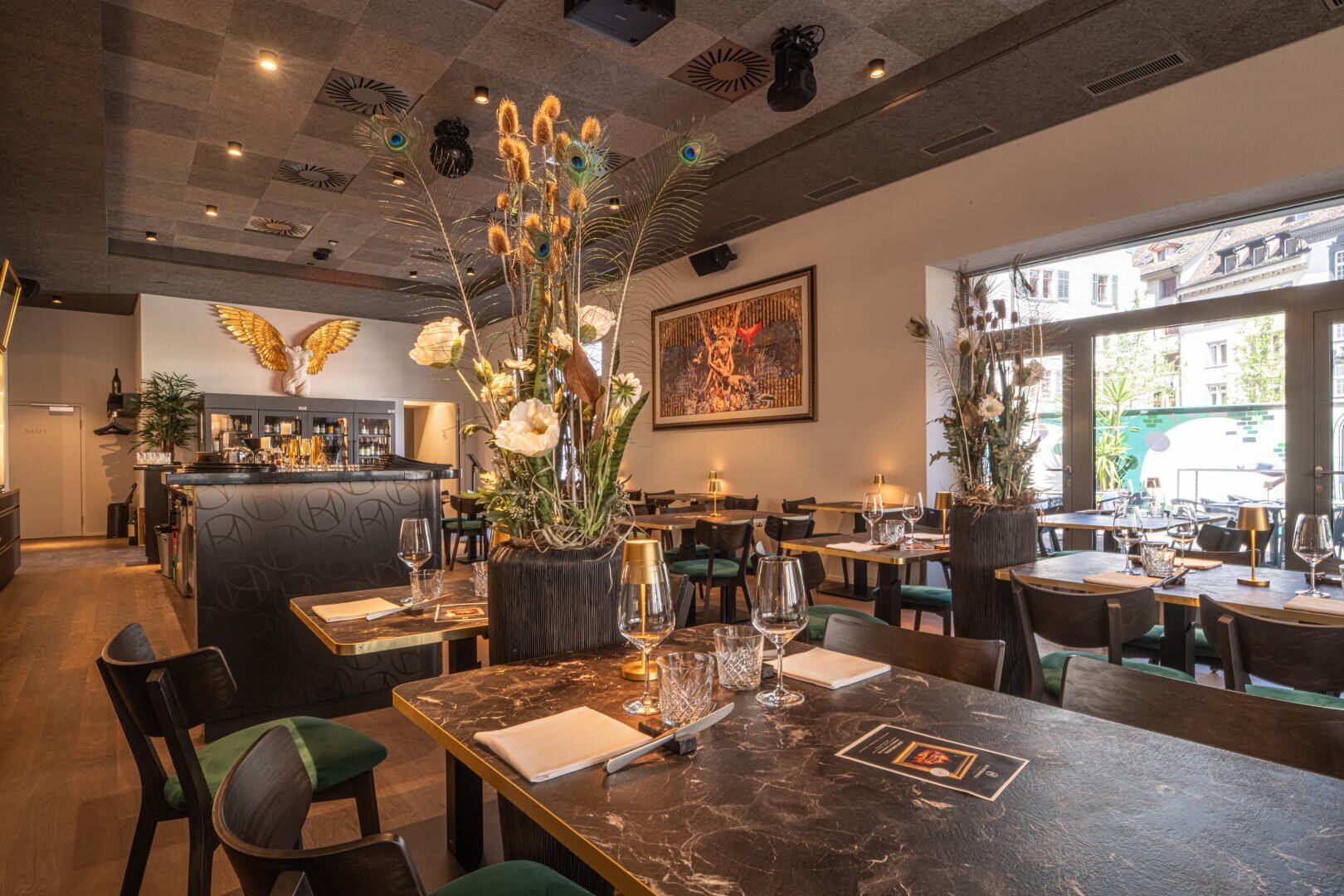 Ein stilvolles, modernes Restaurant mit elegant gedeckten Tischen, großen Blumenkränzen, dunklen Holzmöbeln und einem offenen Blick nach draußen durch Glastüren. Eine Steinadler-Skulptur schmückt den Barbereich.