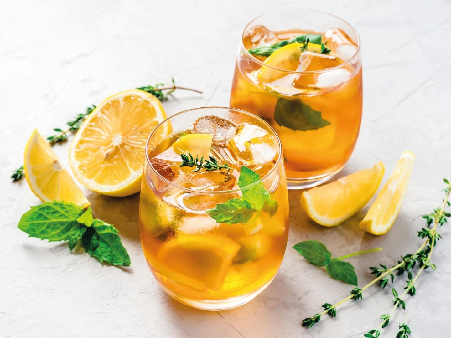 Deux verres de thé glacé, garnis de rondelles de citron, de feuilles de menthe et de brindilles de thym, entourés d'autres rondelles de citron et d'herbes fraîches sur un support clair.