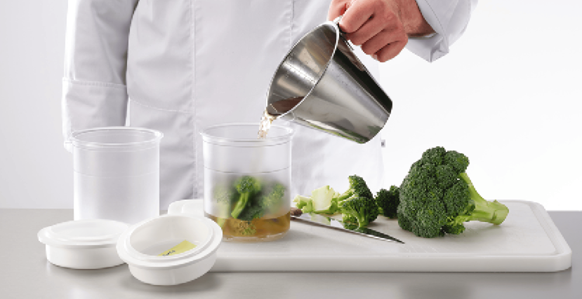 Une personne en tenue de cuisine blanche verse du liquide dans un récipient transparent contenant des brocolis, placé à côté d'une planche à découper avec des brocolis, un couteau et deux récipients à couvercle.