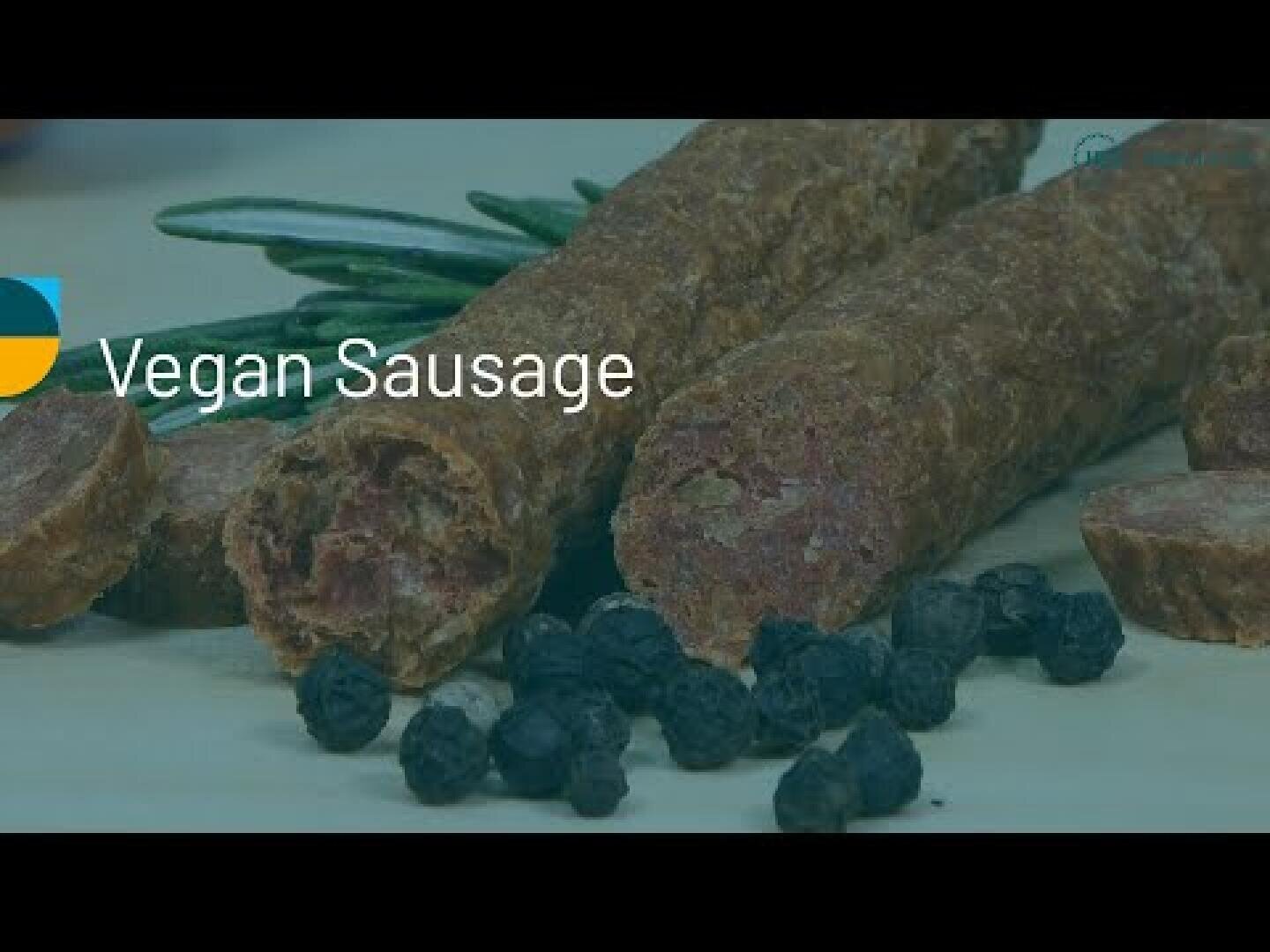Zwei vegane Würste liegen auf einer Holzplatte, eine davon aufgeschnitten. Davor sind ein Kräuterzweig und mehrere dunkle runde Pfefferkörner verstreut. Auf der linken Seite erscheint der Text Vegane Wurst.