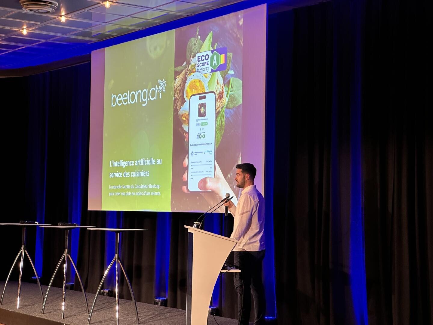 Ein Mann steht an einem Podium und hält einen Vortrag. Hinter ihm ist ein großer Bildschirm zu sehen, auf dem eine Folie mit dem Text "beelonga" und "L'intelligence artificielle au service des cuisiniers" neben einem Mobiltelefon und Bildern von Lebensmitteln zu sehen ist.