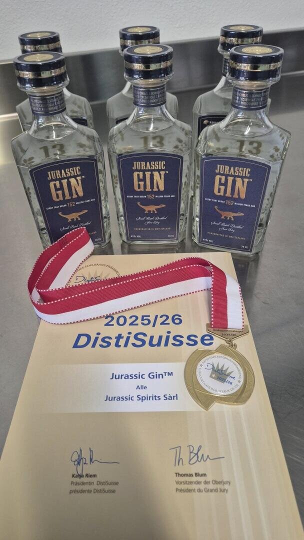 Sechs Flaschen Jurassic Gin stehen auf einer Metallfläche hinter einem DistiSuisse 2025/26-Zertifikat und einer Goldmedaille mit rot-weissem Band, verliehen an Jurassic Spirits Sàrl.