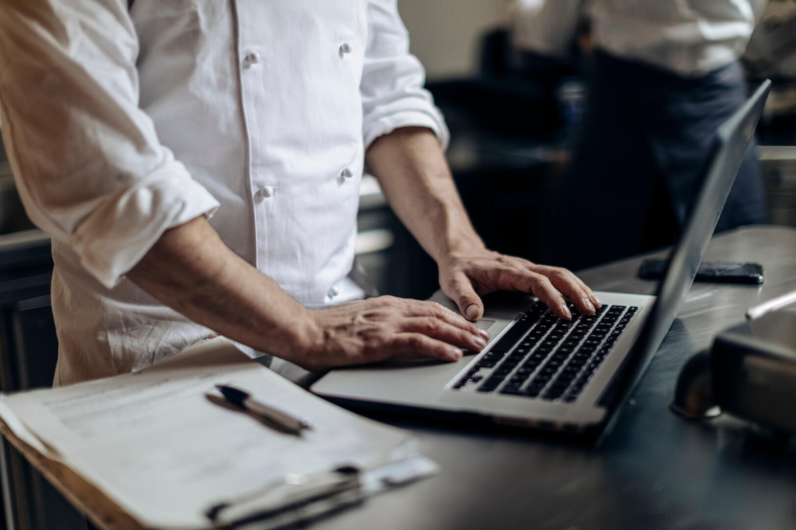 Un cuisinier en uniforme blanc tape sur un ordinateur portable posé sur un comptoir en acier inoxydable, à proximité se trouvent un porte-bloc, un stylo et des papiers dans une cuisine professionnelle.