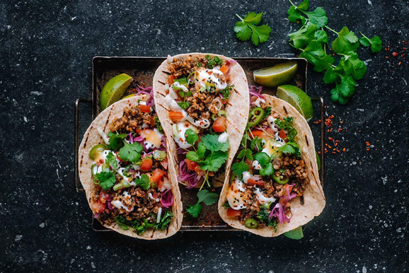 Drei Tacos, gefüllt mit gewürztem veganem Hack aus Mungbohnen, Tomatenwürfeln, violettem Kohl, geschnittenen Jalapeños, frischem Koriander und einer cremigen Sauce, serviert mit Limettenspalten auf einer dunklen Unterlage.