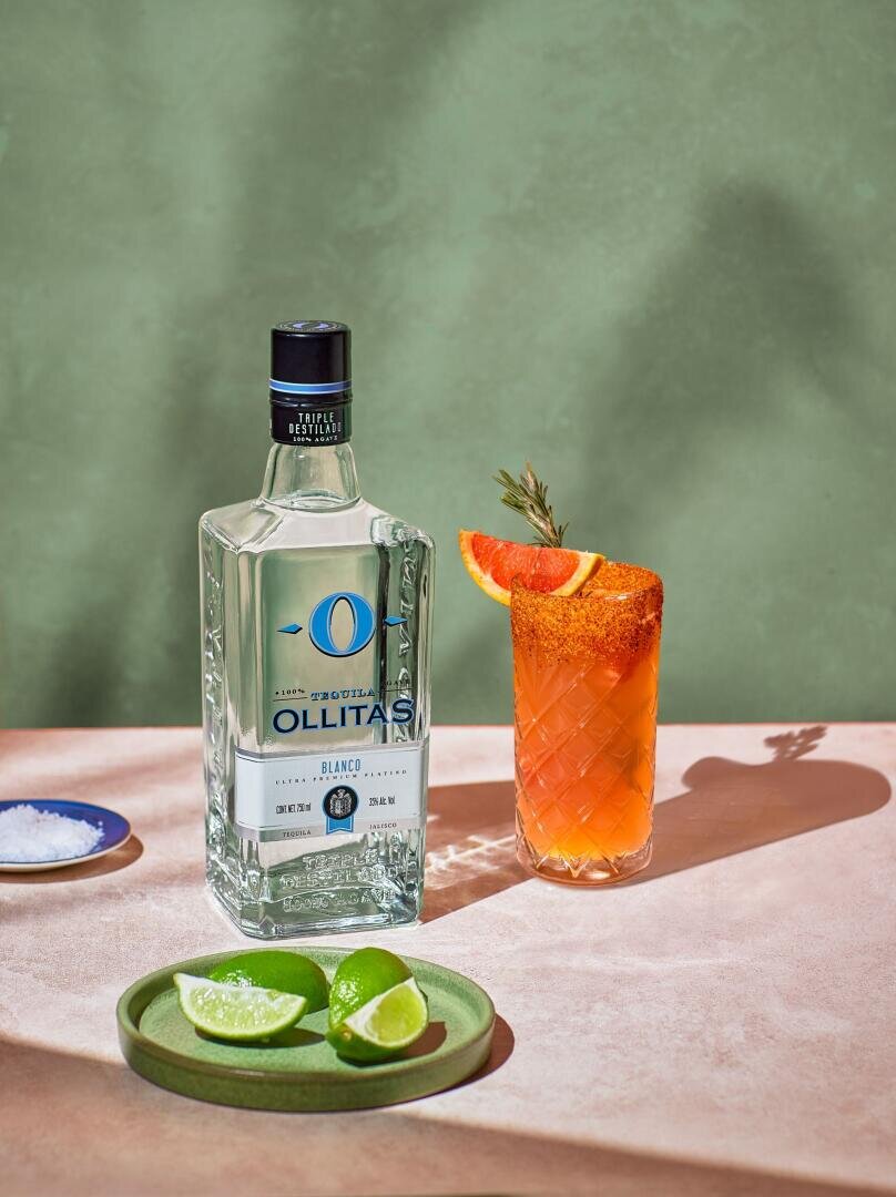 Eine Flasche Ollitas Blanco Tequila steht neben einem mit Grapefruit und Rosmarin garnierten Cocktail, einem Teller mit Limettenspalten und einer kleinen Schale mit Salz auf einer rosafarbenen Fläche vor einem grünen Hintergrund.