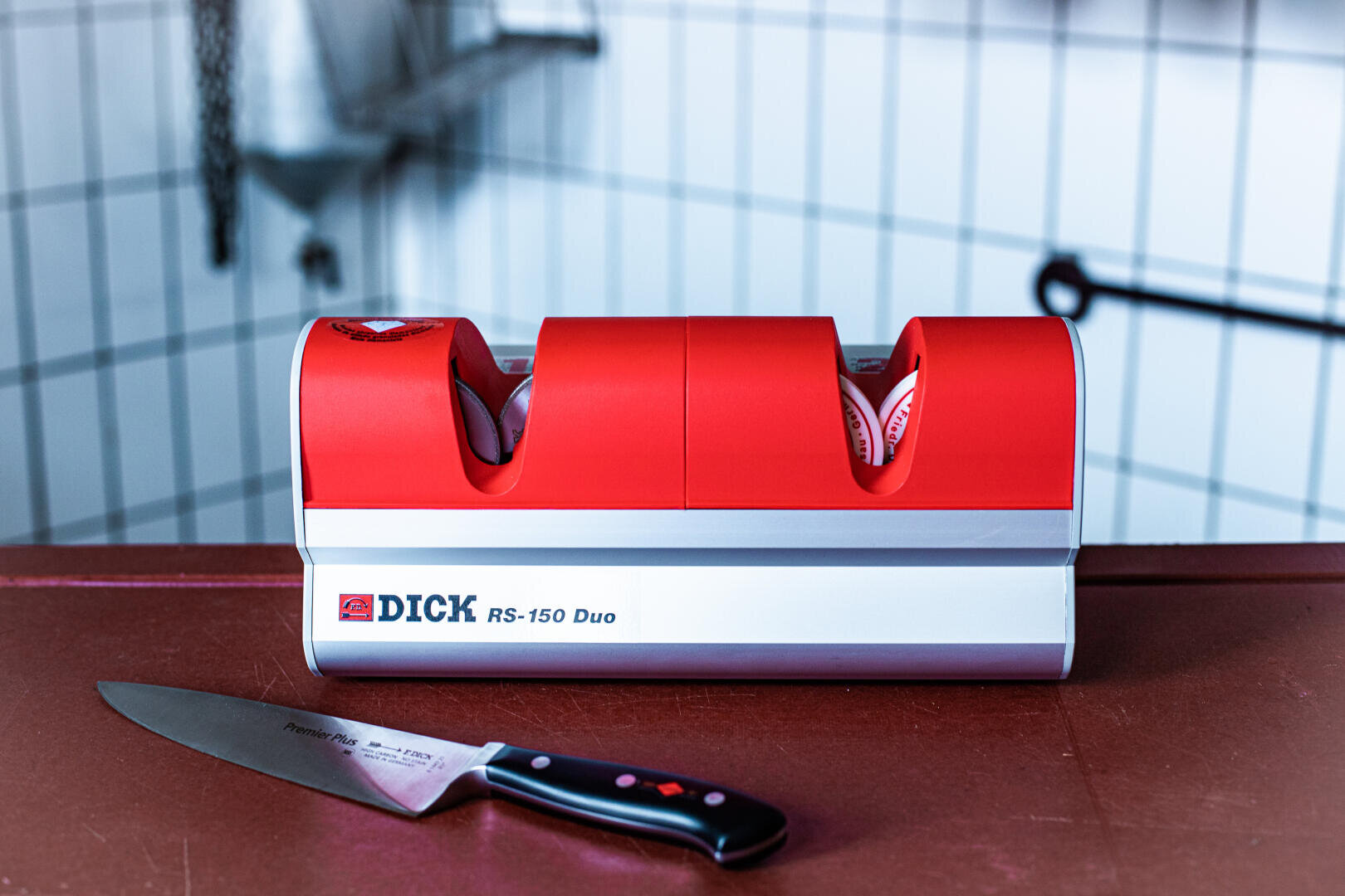 Ein roter und silberner DICK RS-150 Duo-Messerschärfer steht auf einem Tresen, vor dem ein Kochmesser liegt. Im Hintergrund sind weiß geflieste Wände und ein Metallregal zu sehen.