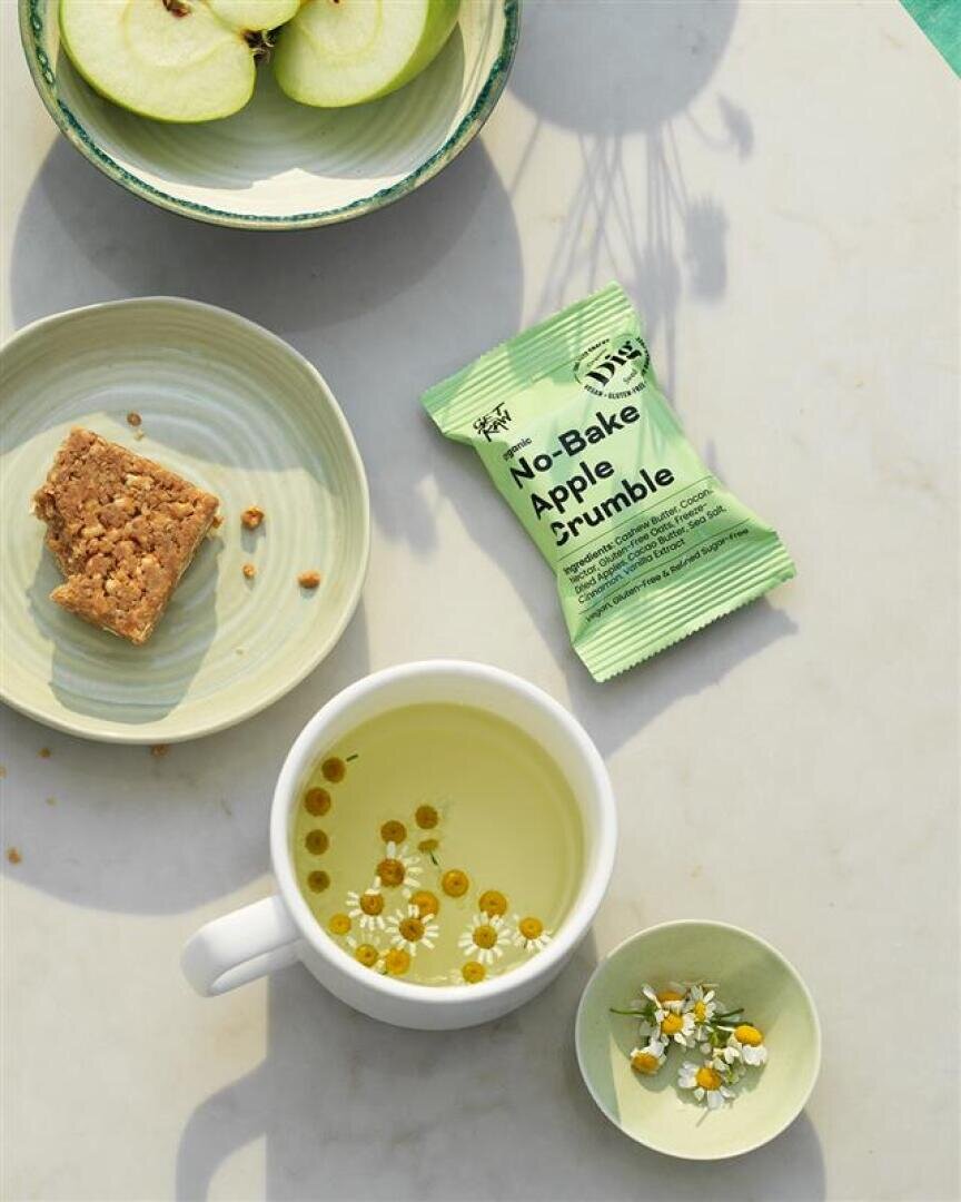 Eine Tasse Kamillentee mit frischen Blüten, eine Schale mit in Scheiben geschnittenen grünen Äpfeln, ein Granola-Quadrat auf einem Teller und ein verpackter Apfelstreusel-Snack auf einer weißen Fläche.