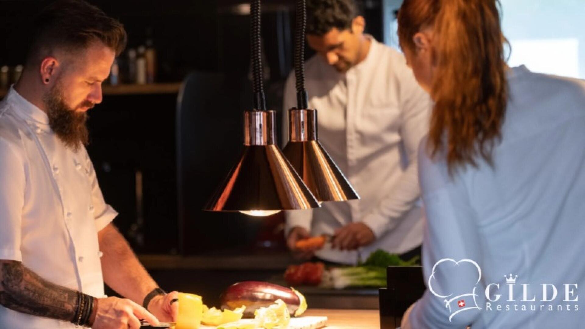 Drei Köche in weißen Uniformen bereiten frisches Gemüse in einer modernen, sanft beleuchteten Küche zu. In der unteren rechten Ecke ist das Logo von Gilde Restaurants zu sehen.