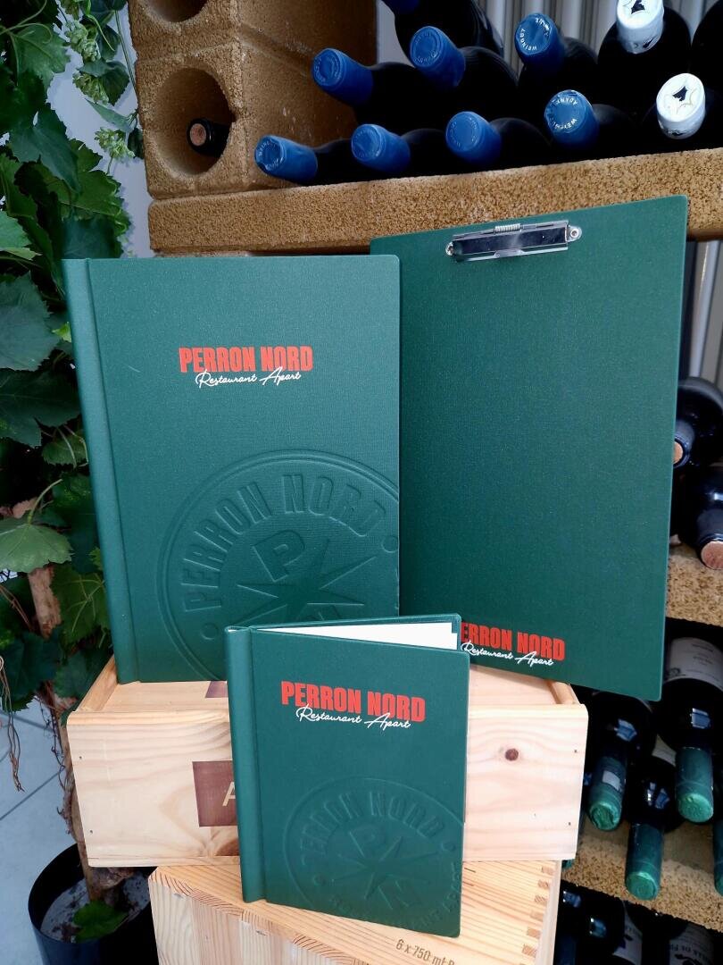 Drei grüne Speisekarten für das Perron Nord Ristorante Agnese sind auf einer Holzkiste ausgestellt, mit Weinflaschen und Weinreben im Hintergrund. Die Speisekarten sind unterschiedlich groß und mit orangefarbener und weißer Schrift versehen.