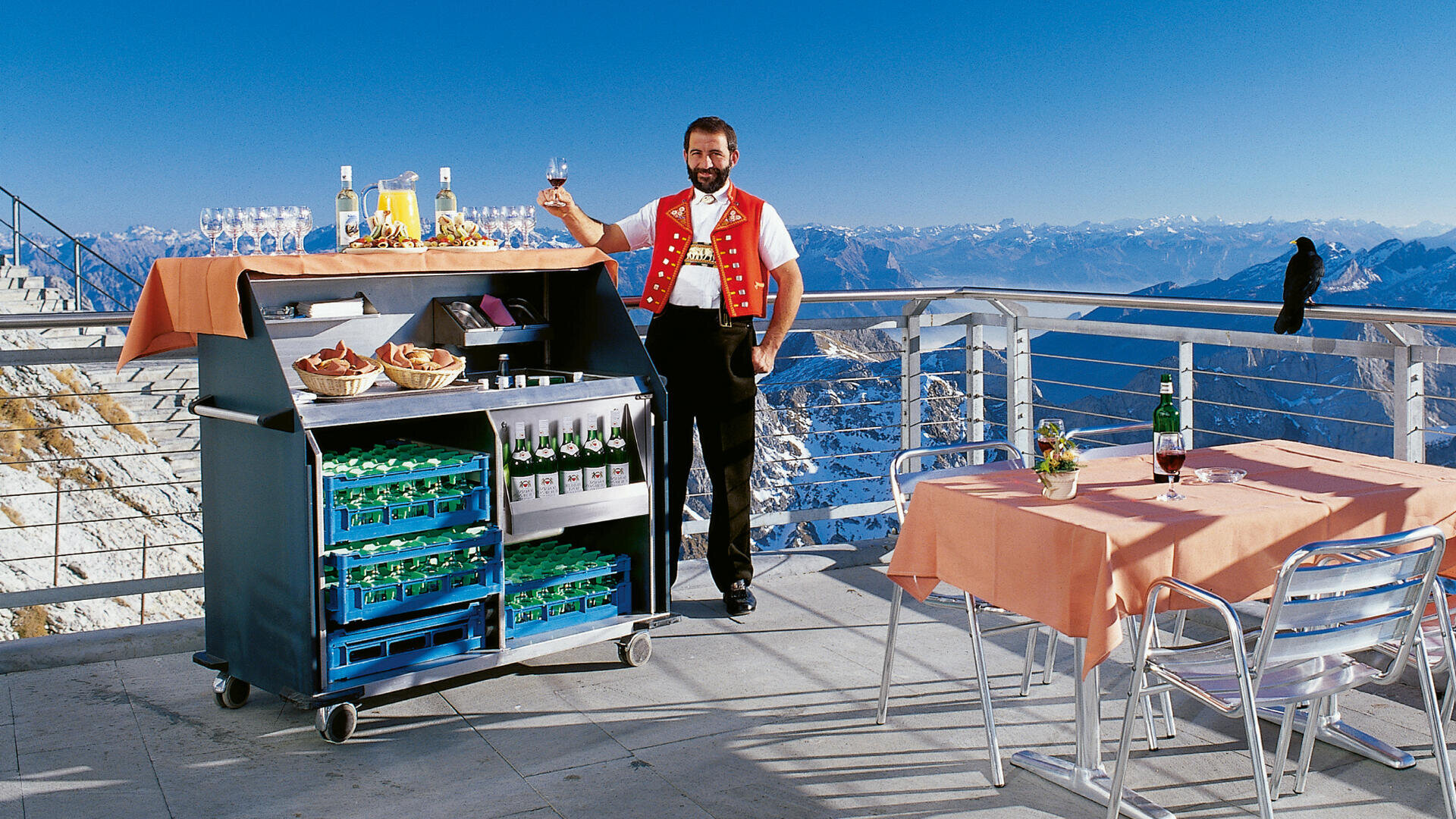 Un homme en gilet rouge se tient debout, un verre à la main, à côté d'un chariot de boissons approvisionné, sur une terrasse de montagne. Il y a des tables recouvertes de draps orange et des montagnes enneigées en arrière-plan sous un ciel bleu limpide.