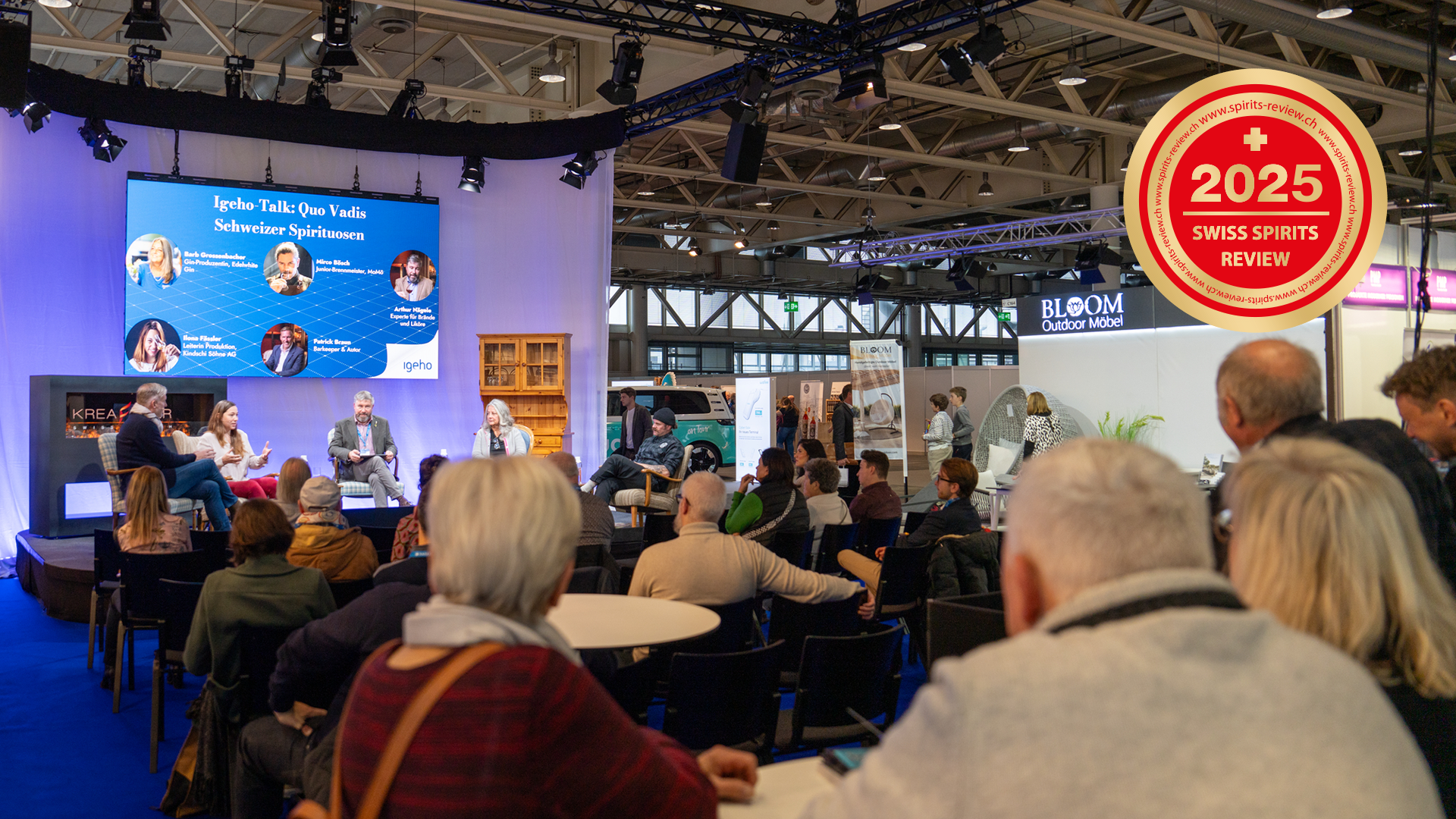 Une table ronde se déroule sur une scène dans un hall et est suivie par un public. Des informations sur les orateurs sont affichées sur un grand écran. Dans le coin supérieur droit, un badge rouge de la 2025 Swiss Spirits Review est affiché.