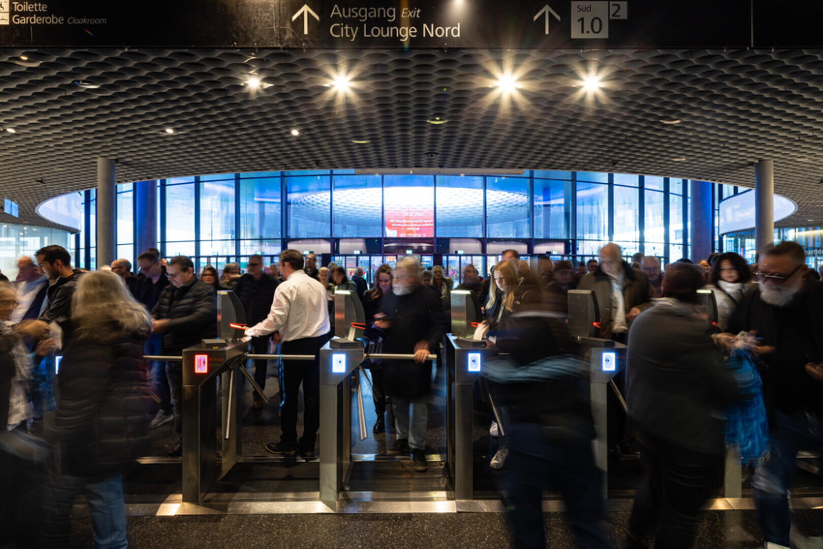 Eine geschäftige Menschenmenge passiert die Fahrkartenschranken eines modernen Bahnhofs, mit beleuchteten Schildern und einem großen Glaseingang im Hintergrund.