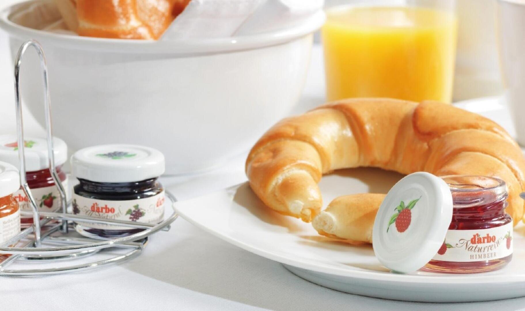 Un croissant sur une assiette blanche à côté d'un petit pot de confiture de framboise, de mini-verres contenant d'autres confitures, d'une corbeille à pain et d'un verre de jus d'orange sur une nappe blanche.