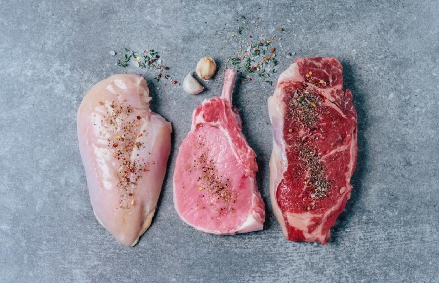Drei rohe Fleischstücke - eine Hühnerbrust, ein Schweinekotelett und ein Rindersteak - sind auf einer grauen Fläche angeordnet und mit Gewürzen, Kräutern und Knoblauchzehen bestreut.