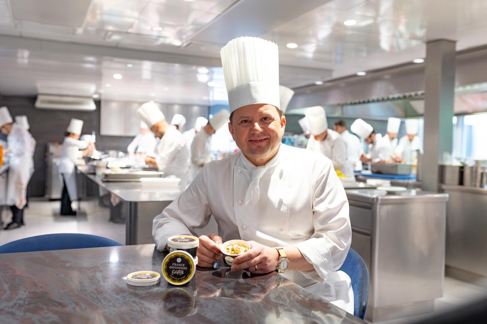 Ein Koch in weißer Uniform und großer Mütze sitzt an einem Tisch in einer professionellen Küche und hält eine Schüssel mit Essen in der Hand. Andere Köche arbeiten im Hintergrund. Auf dem Tisch vor ihm stehen zwei Behälter mit Kaviar.
