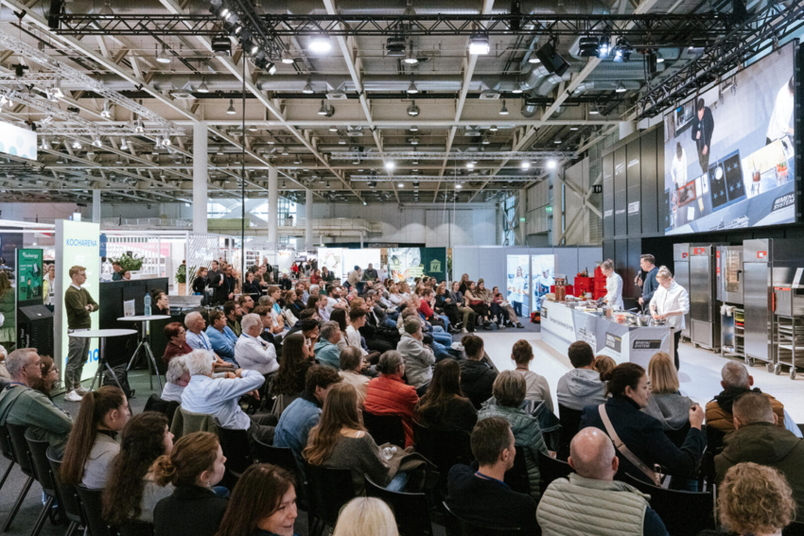 Ein großes Publikum sitzt in Reihen und verfolgt eine Live-Kochvorführung auf der Bühne einer belebten Veranstaltung oder Messe, bei der Köche und Moderatoren an Theken unter hellen Lichtern arbeiten.
