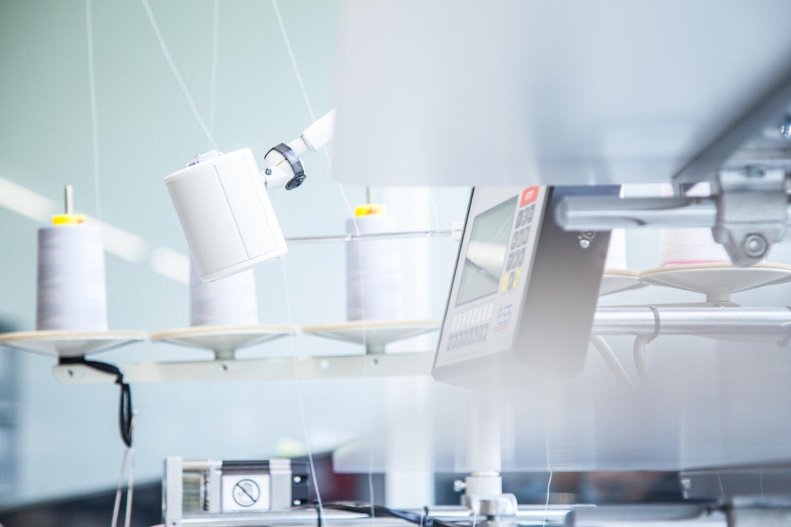 Nahaufnahme von weißen Garnspulen auf einer Maschine in einer modernen Textilfabrik. Ein digitales Bedienfeld und ein Roboterarm sind zu sehen, die eine saubere, hochtechnologische Produktionsumgebung schaffen.