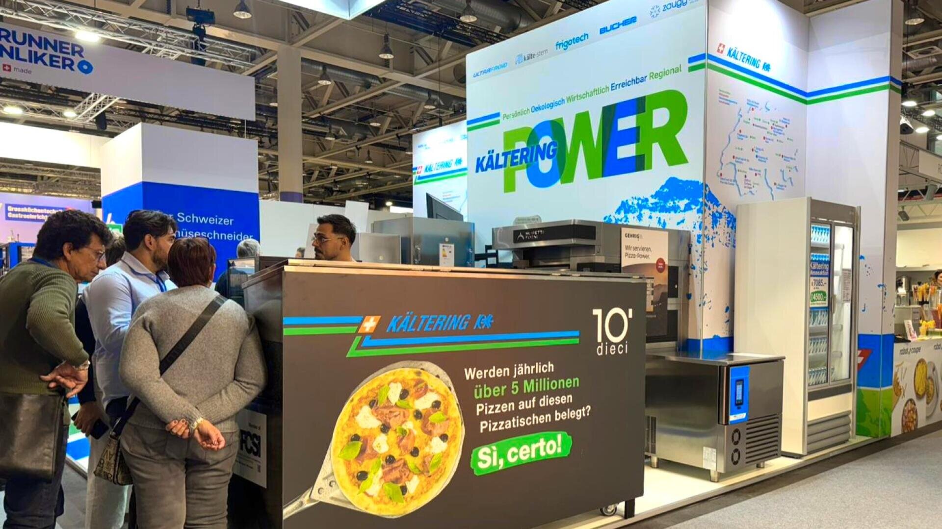 Auf einer Messe stehen Menschen an einem Pizzastand, der mit einem großen Display für Pizza und dem Slogan "Si, certo!" wirbt. Der Stand ist mit Küchengeräten und einem bunten Schild mit einer Landkarte und dem Wort "KÄLTERING" in blauer Schrift ausgestattet.