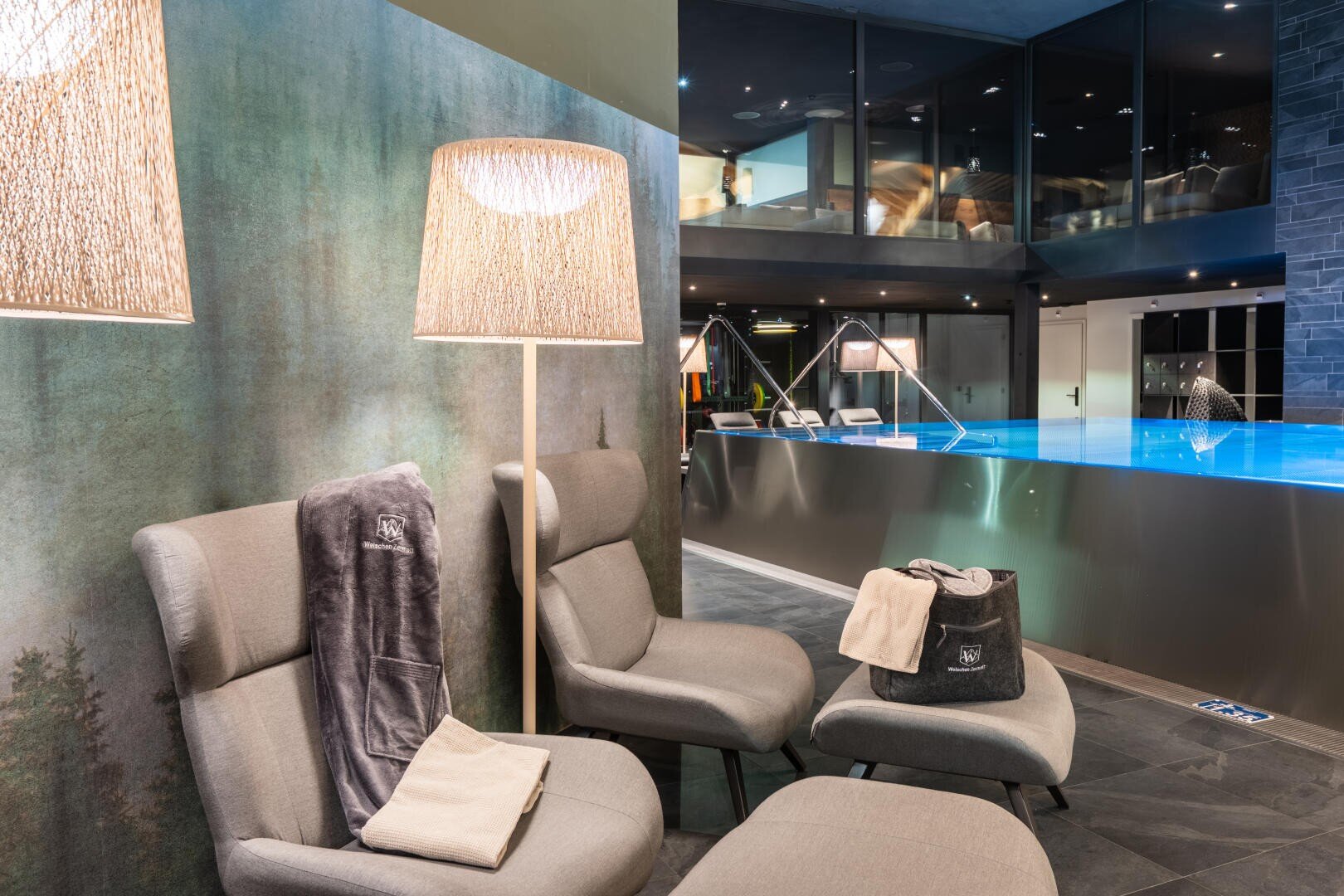 Moderne Indoor-Spa-Lounge mit drei grauen Stühlen, einer Stehlampe, Handtüchern und einem Pool mit blauem Wasser. Der Raum bietet große Fenster, sanfte Beleuchtung und eine ruhige, entspannende Atmosphäre.