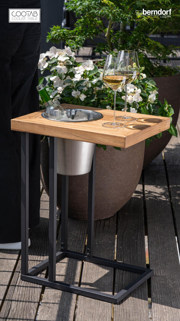 Une petite table en bois avec un seau à glace en métal intégré contenant une bouteille de vin, deux verres à vin remplis sur le dessus, et des fleurs blanches en pot à l'arrière-plan sur un patio en bois.
