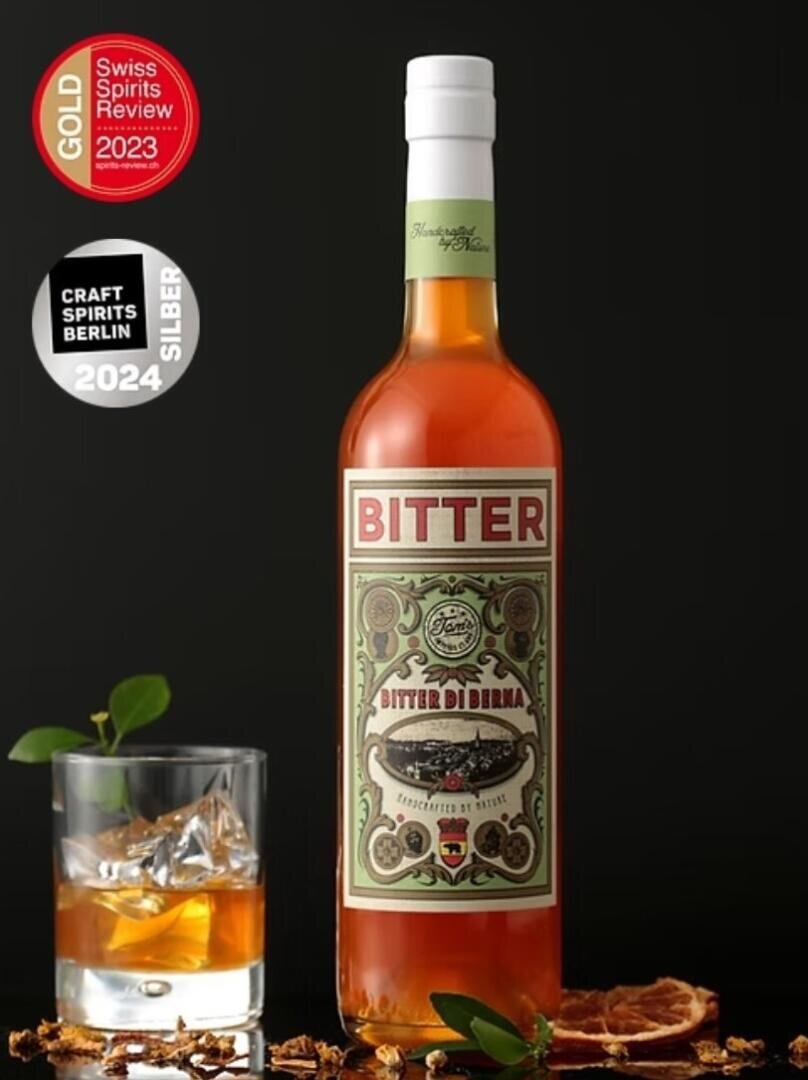 Eine Flasche Bitter Di Berna mit verschnörkeltem Etikett steht neben einem Glas Whiskey mit Eis und einer Zitrusgarnitur; abgebildet sind die Auszeichnungsplaketten der Schweizerischen Spirituosenschau 2023 (Gold) und 2024 Craft Spirits Berlin (Silber).