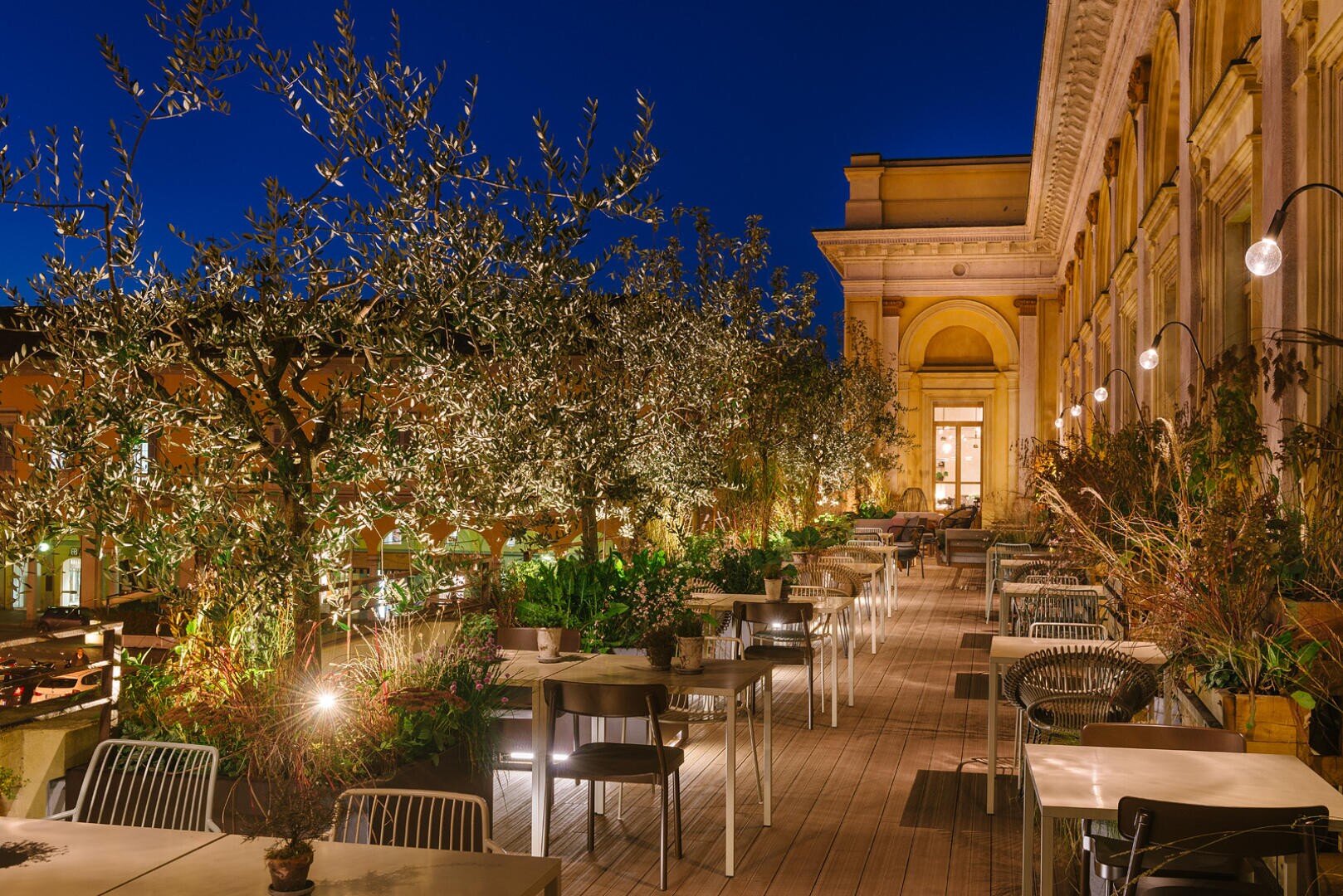 Élégante terrasse extérieure de nuit avec des tables et des chaises bien disposées, entourée d'oliviers et de plantes, ornée de guirlandes lumineuses. L'architecture classique et l'éclairage chaleureux créent une atmosphère douillette et accueillante.