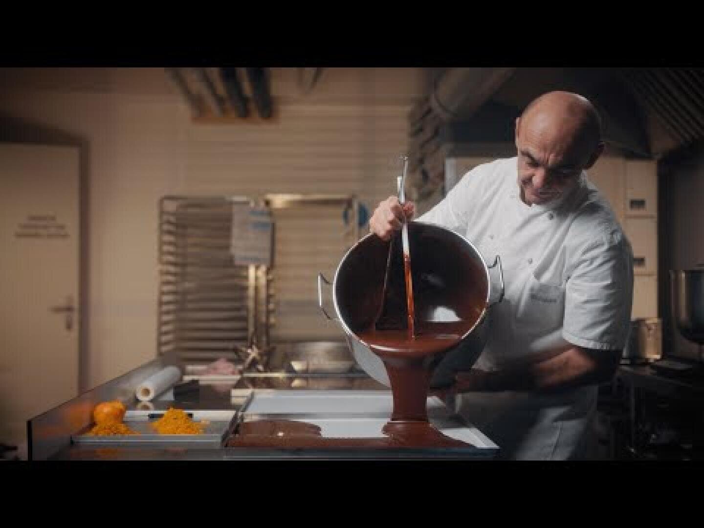 Un chef en uniforme blanc verse du chocolat fondu d'un grand bol sur un plateau dans une cuisine commerciale, avec des oranges et des équipements de cuisine visibles à l'arrière-plan.
