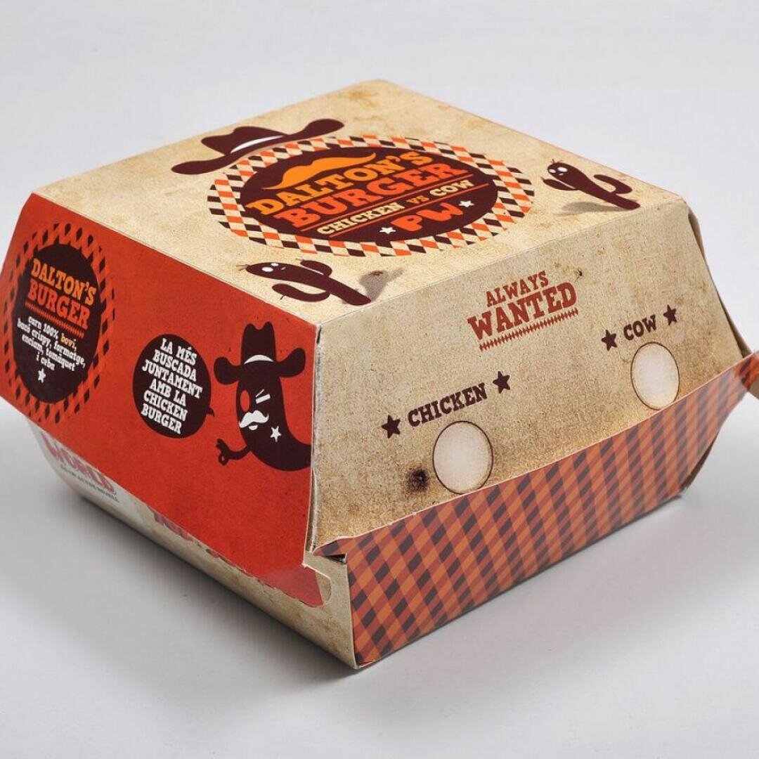 Eine Burger-Box zum Mitnehmen mit einem Western-Design, das eine Cowboy-Silhouette, Kakteen und den Text "Daltons Burger" und "Always Wanted Chicken or Cow" mit Kästchen für Huhn und Kuh zeigt.